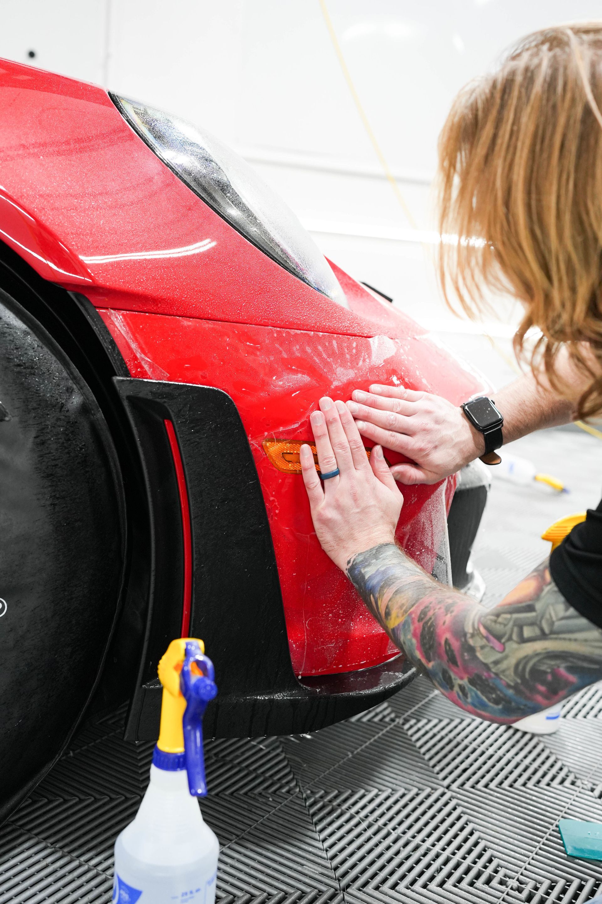Person applying film to a red car's fender. They are using their hands, indoors.
