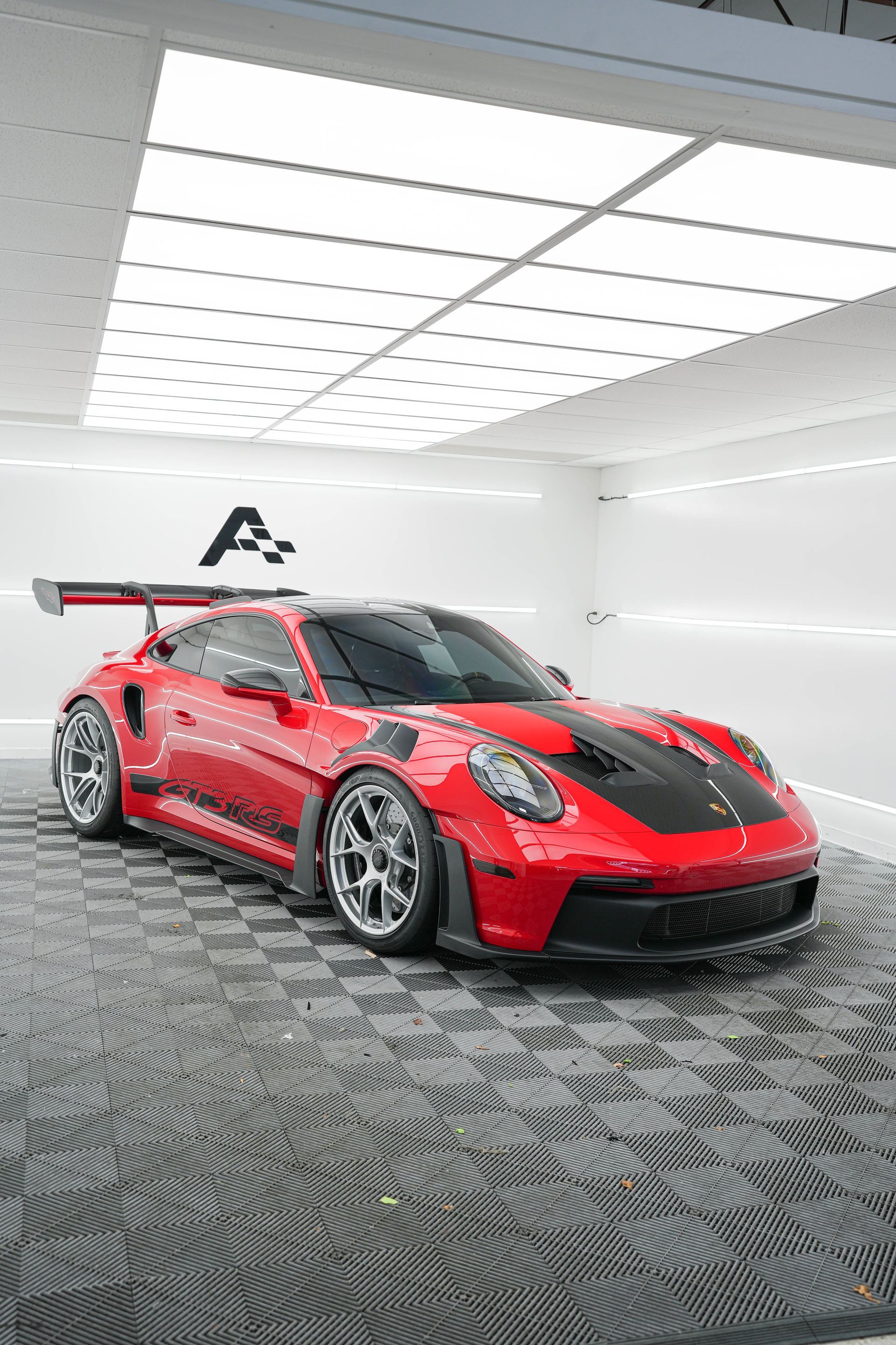 Red Porsche 911 GT3 RS in a clean, well-lit garage with black stripes on the hood.