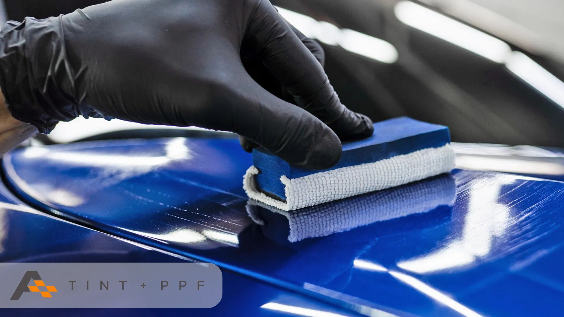 A gloved hand applying a ceramic coating to the surface of a shiny blue car using an applicator bloc