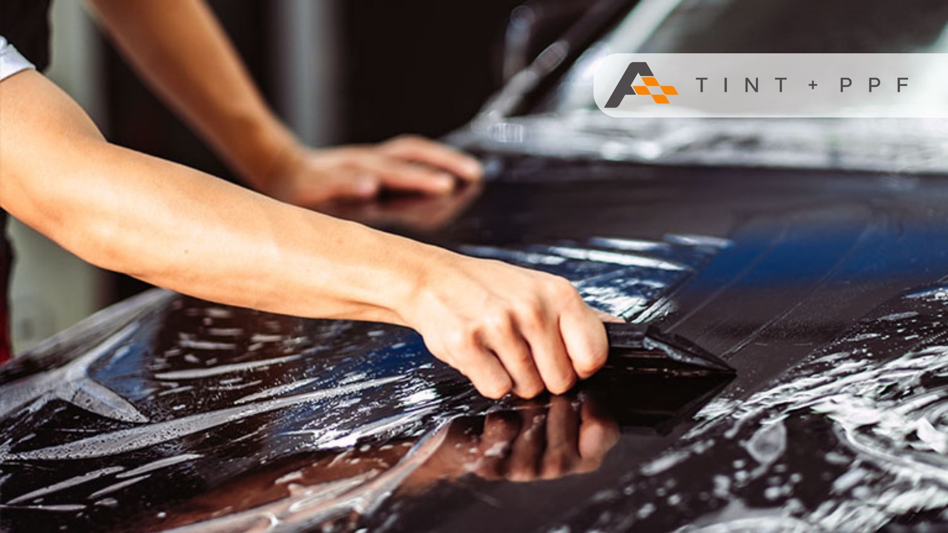 A technician using a squeegee to apply paint protection film (PPF) to a black car, ensuring even cov