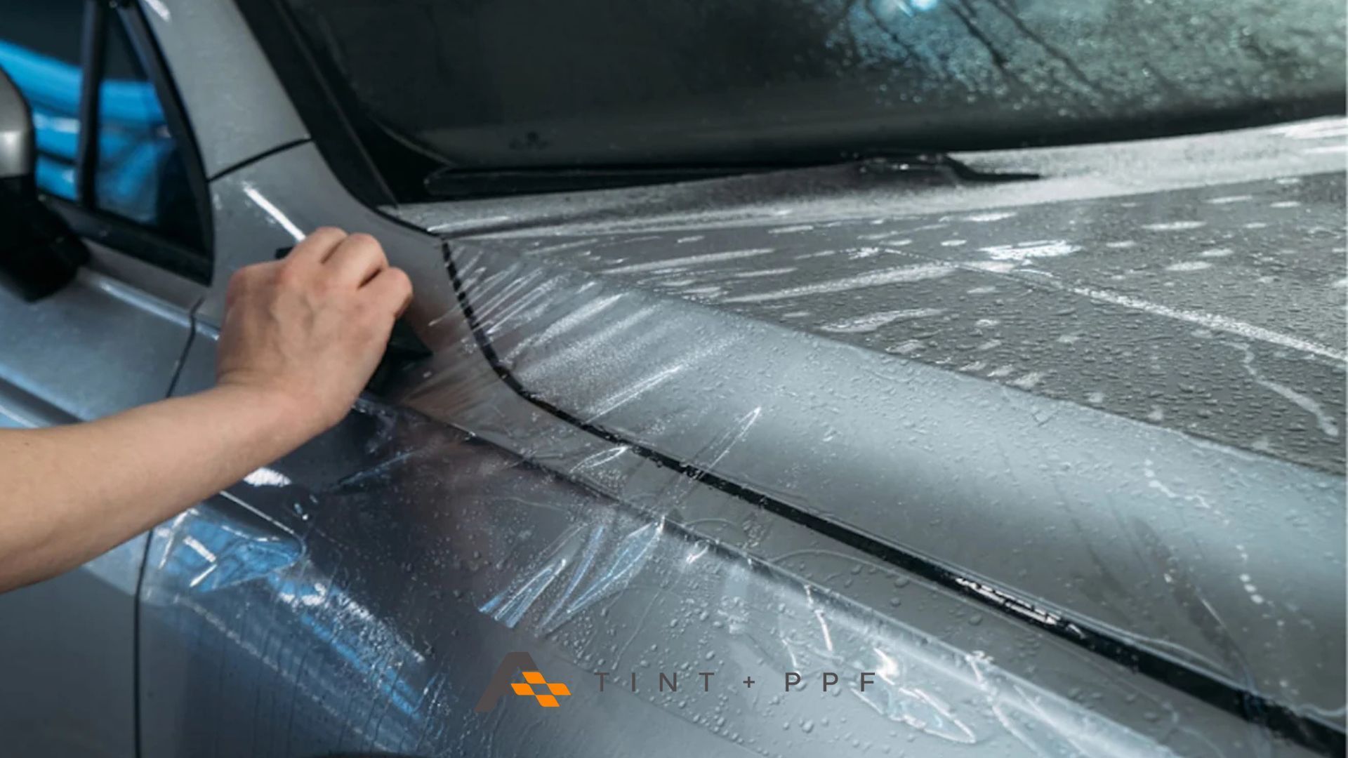 A close-up of a gloved hand smoothing a transparent film onto a silver car fender.