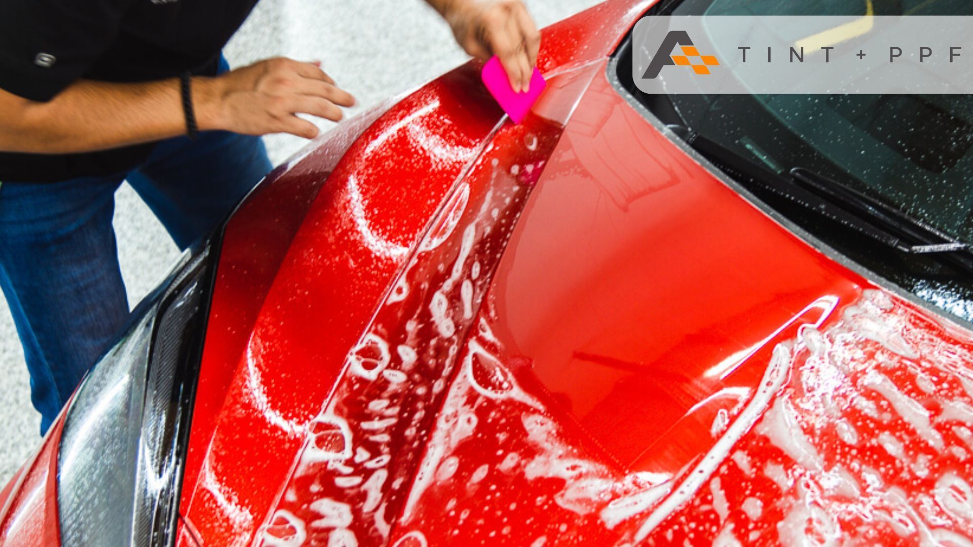 A person applies a protective film to the hood of a red car using a pink squeegee, with soapy water 