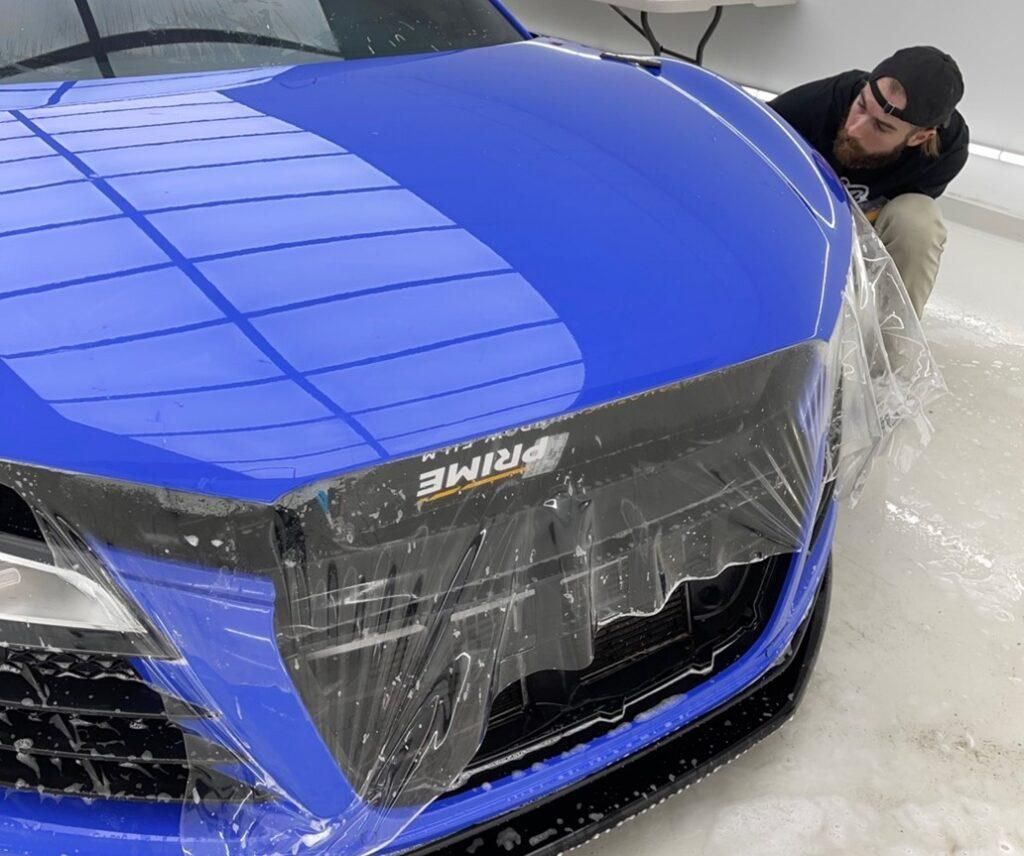 A man is covering the front of a blue car with plastic wrap.
