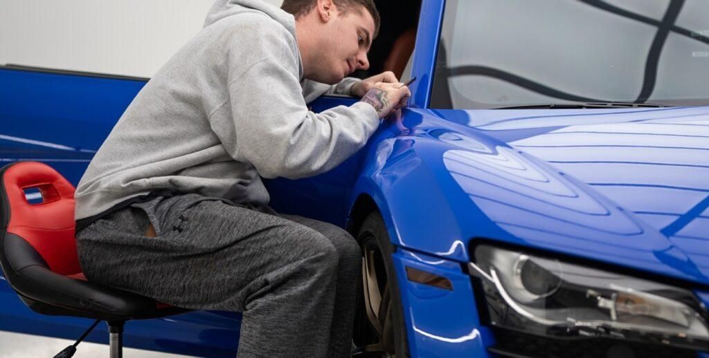A man is sitting on a stool working on a blue car.