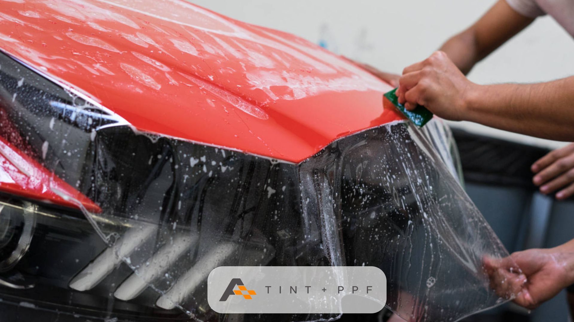 A red car hood with a technician applying a clear protective film using a green squeegee. The wet su