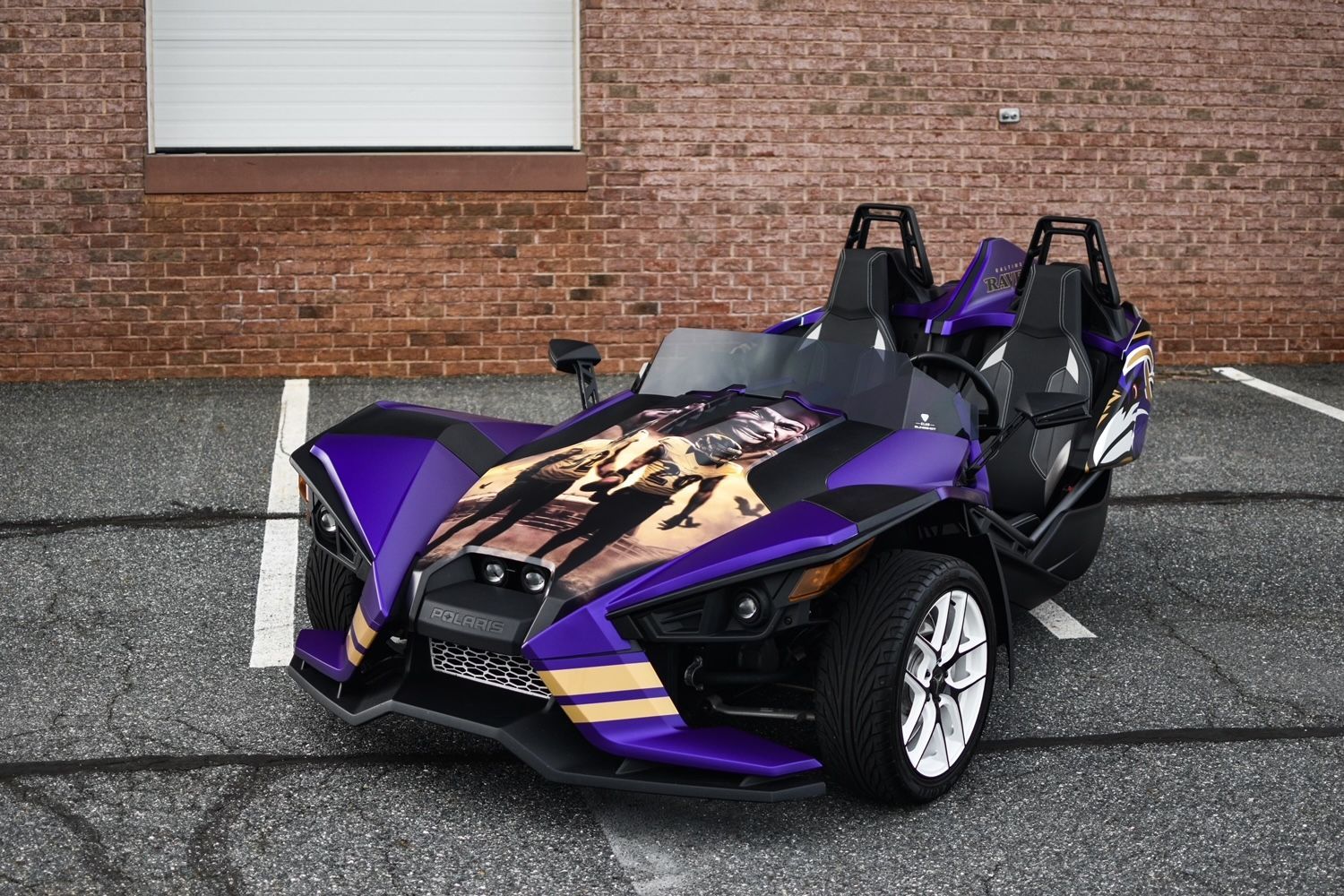 A purple slingshot is parked in a parking lot in front of a brick building.