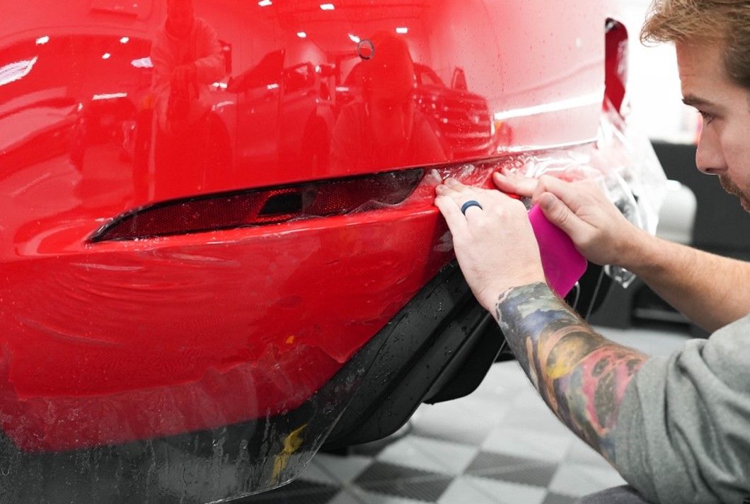 A man is wrapping the rear bumper of a red car.