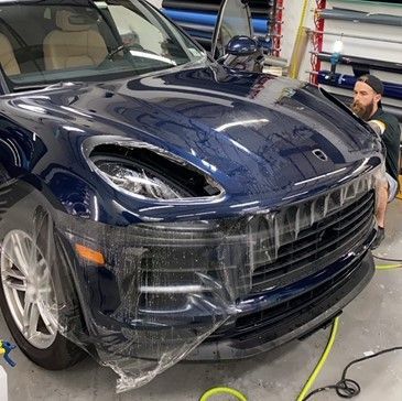 A man is standing next to a blue porsche macan turbo wrapped in plastic.