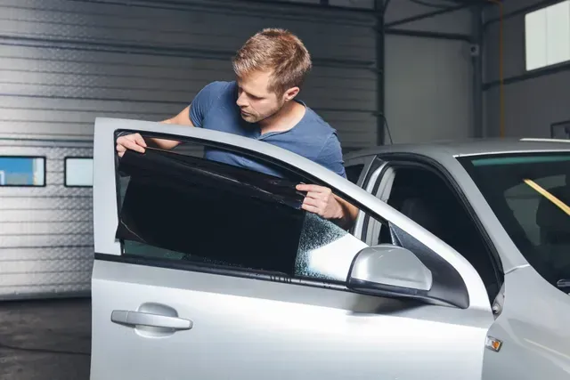 A man carefully applies dark window tint film to the front side window of a silver car inside a gara