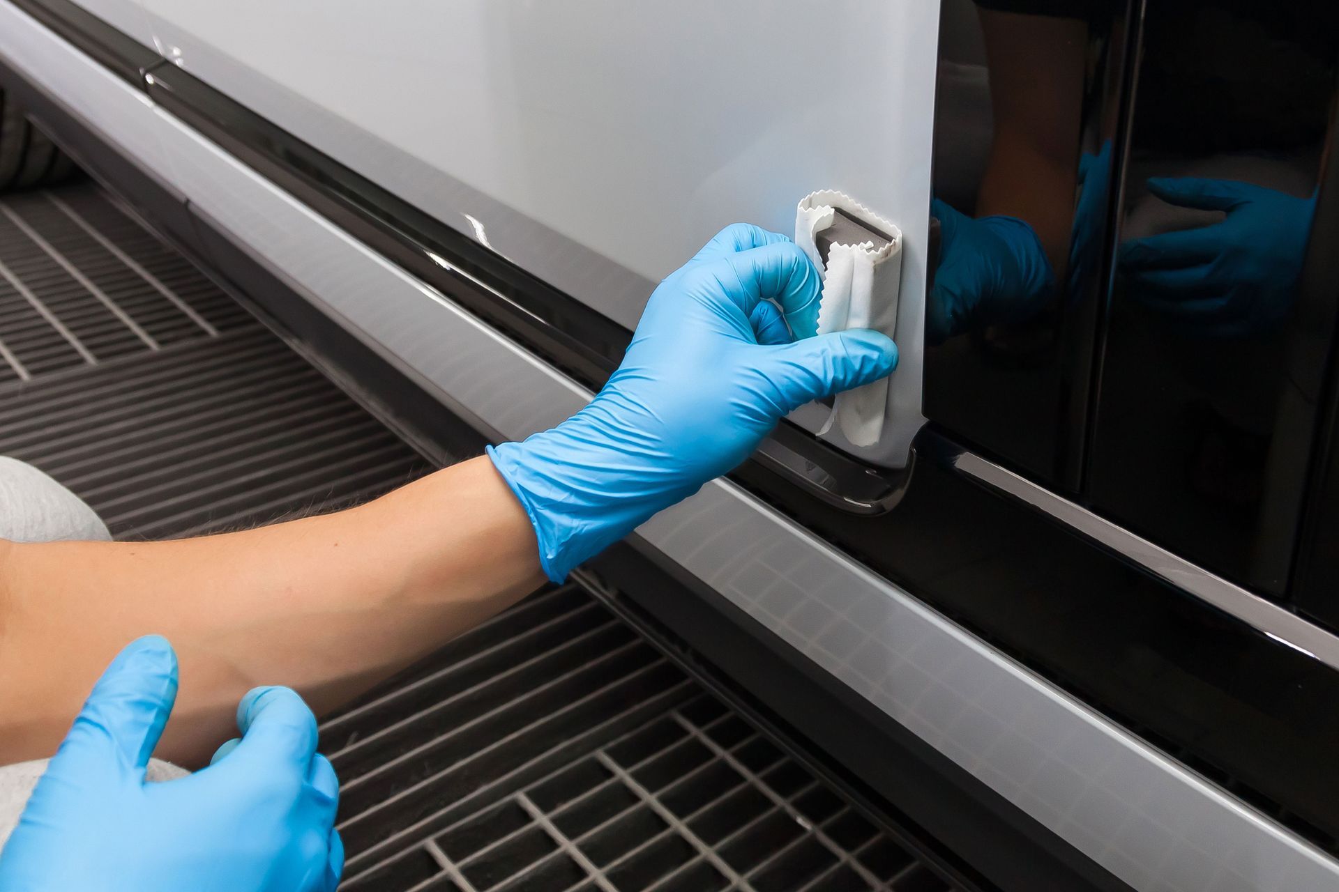 Person in blue gloves using a tool on a car door. Black and silver car in a garage.