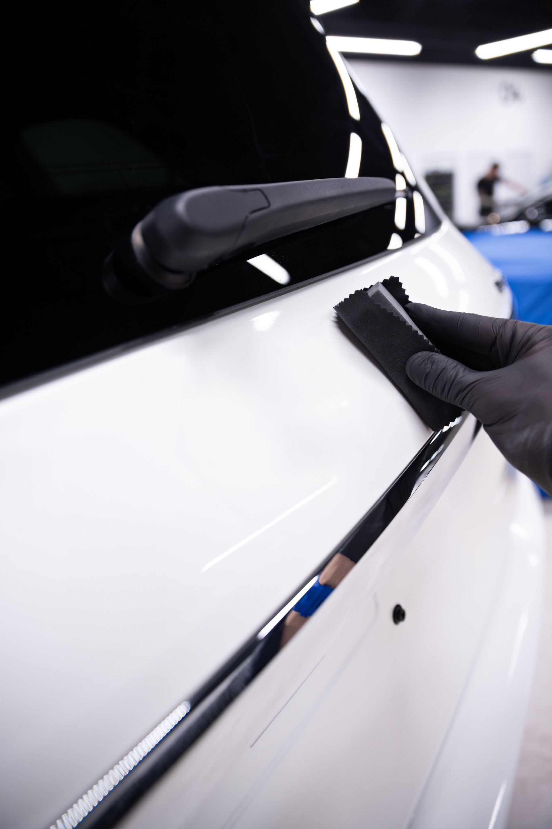 A gloved hand applies a black coating to a white car's rear panel near the window.