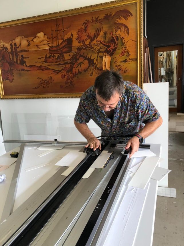 A Man Is Working On A Machine In Front Of A Large Painting — The Picture Framer In Parramatta Park, QLD
