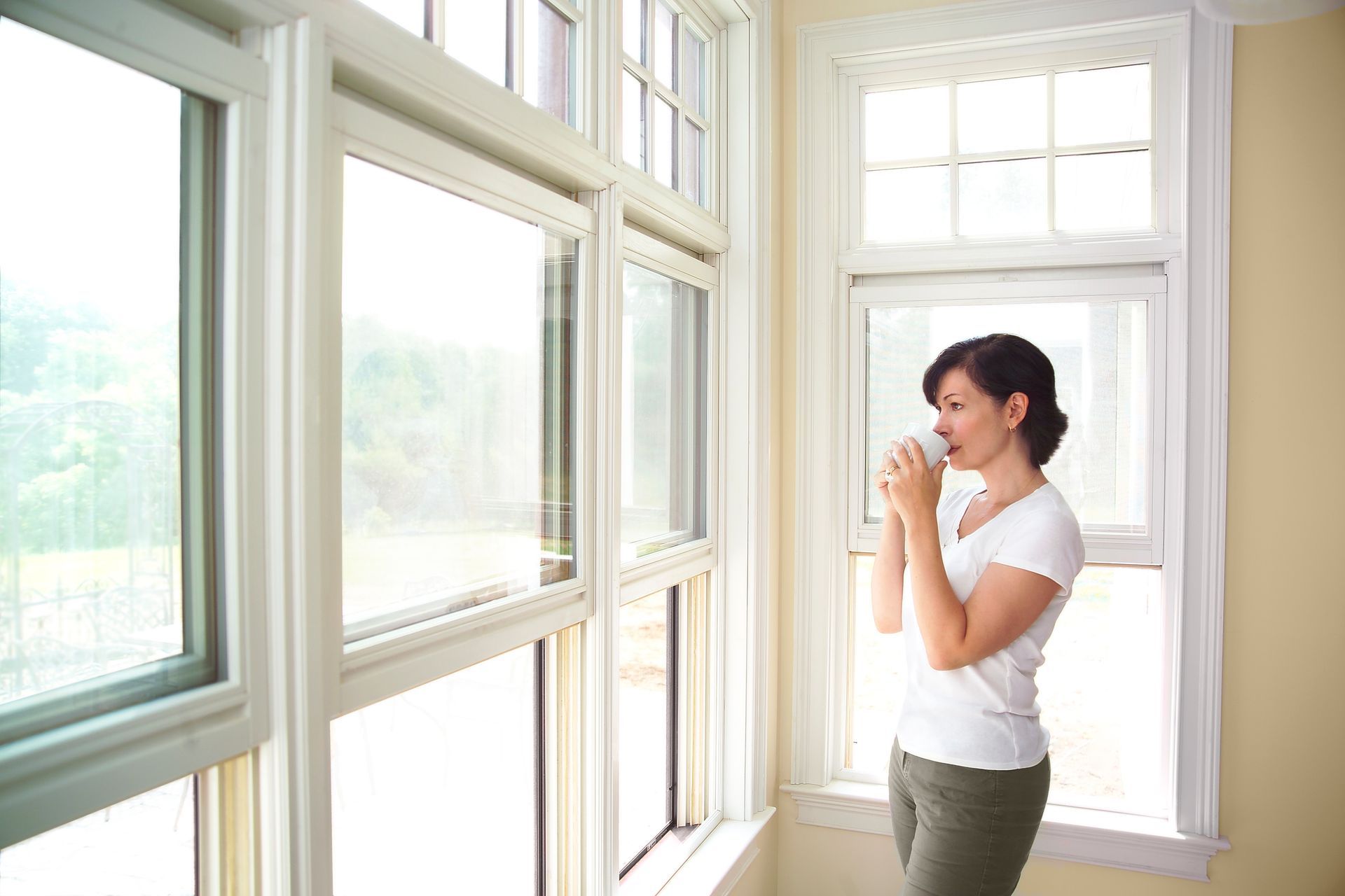 A woman holding a mug is looking out her newly installed windows. A woman holding a mug is looking out her newly installed windows.
