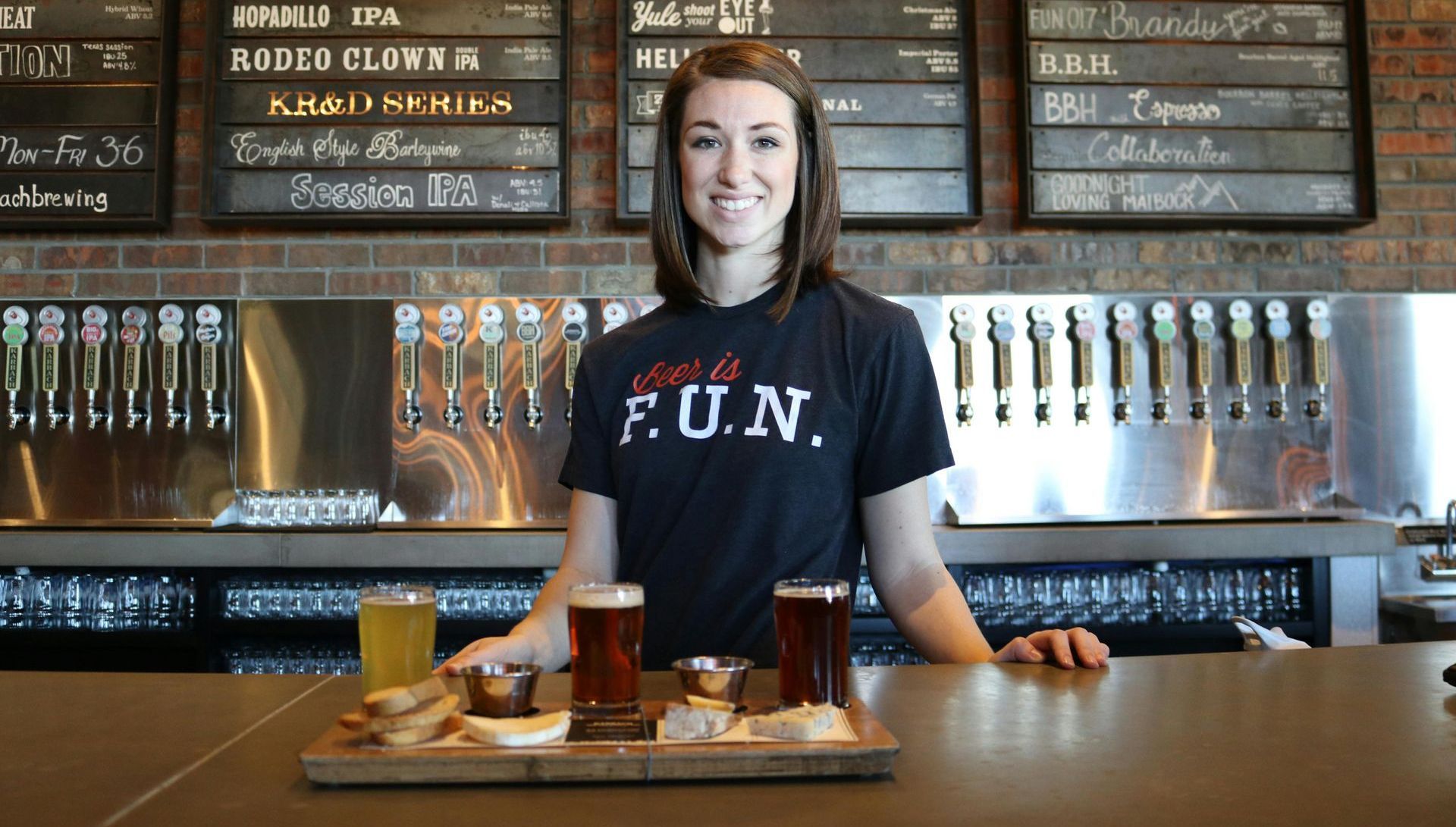 A woman wearing a shirt that says fun is standing at a bar holding a tray of beer.