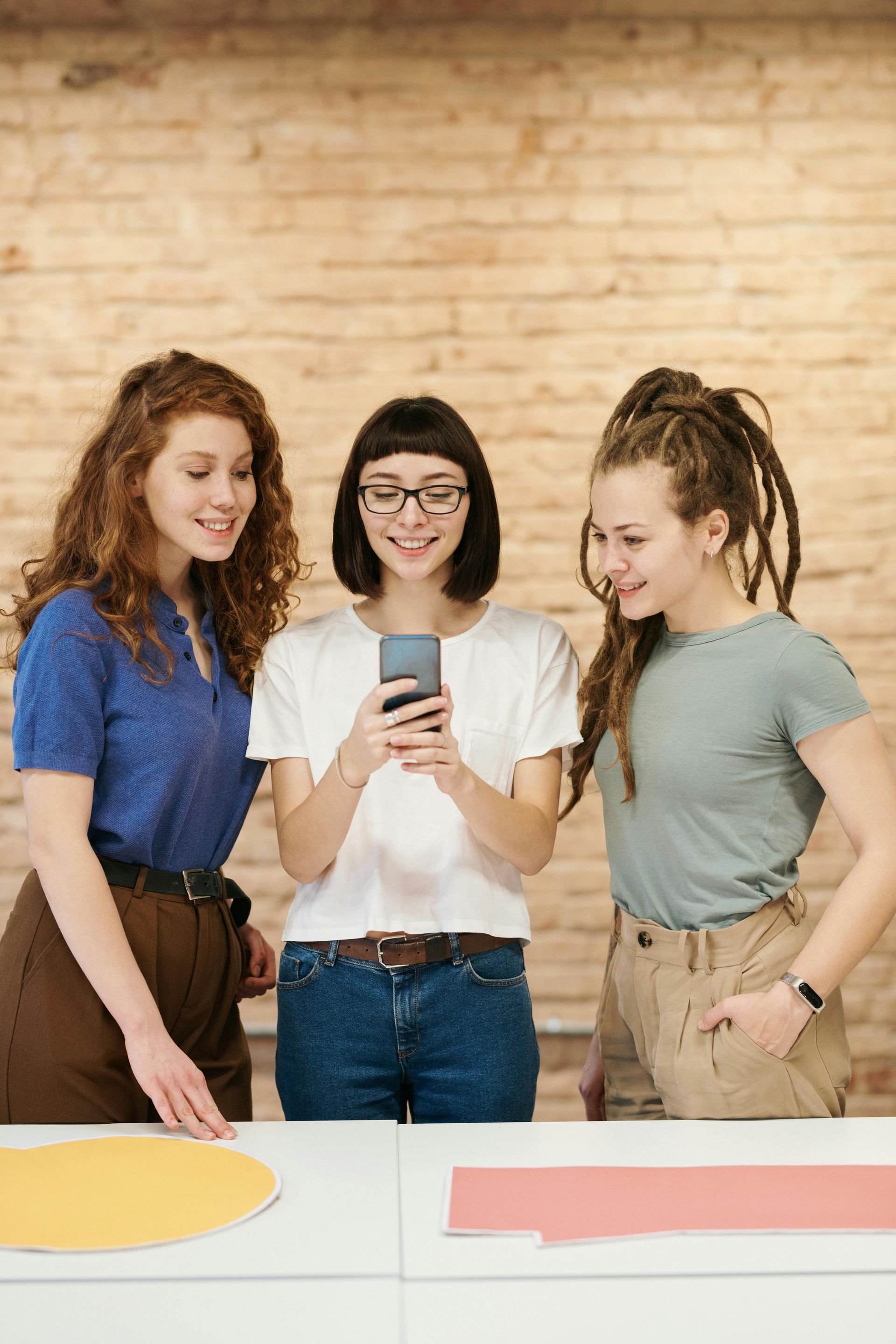 Three women are standing around a table looking at a cell phone.