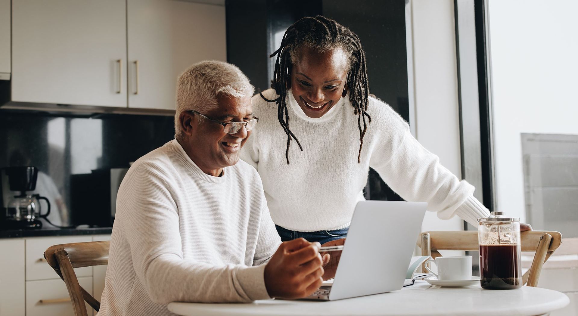 An older Black man and younger woman smile at a laptop in a kitchen. Coffee and a cup are on the table.