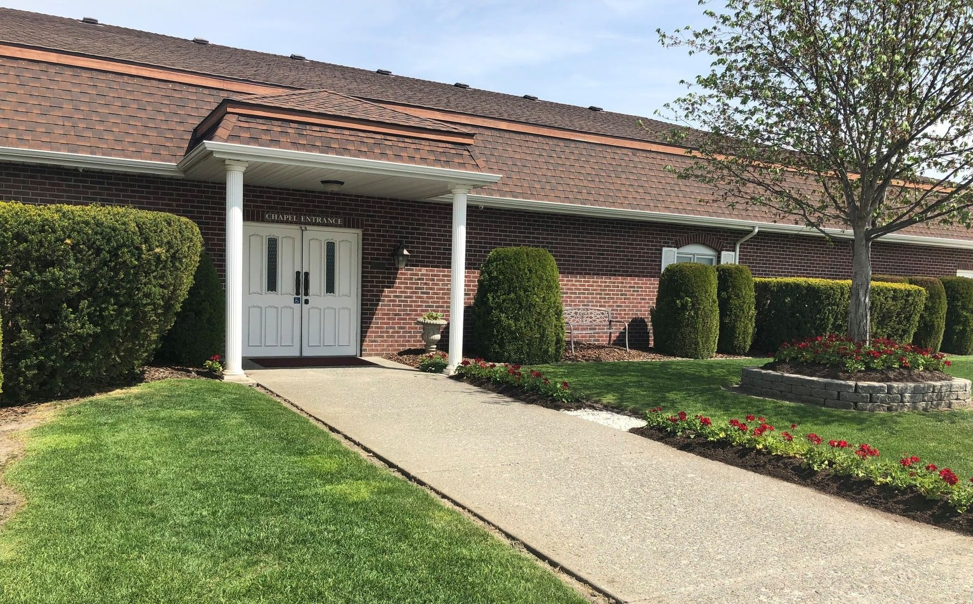 Low brick building with white doors, a concrete walkway, and green landscaping.