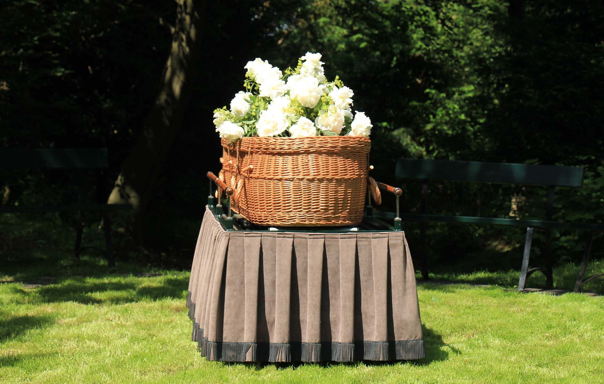 Wicker coffin with white flowers on a pleated skirt-covered stand in a grassy outdoor setting.