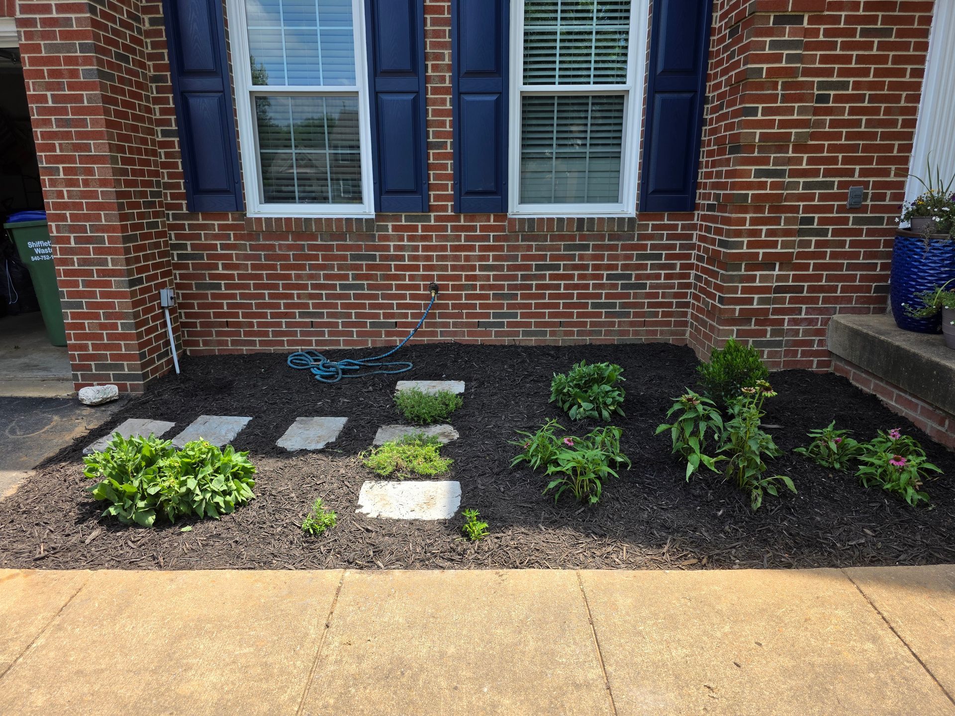 Fresh mulch landscaping along a brick house with small plants and a blue yard sign near the walkway