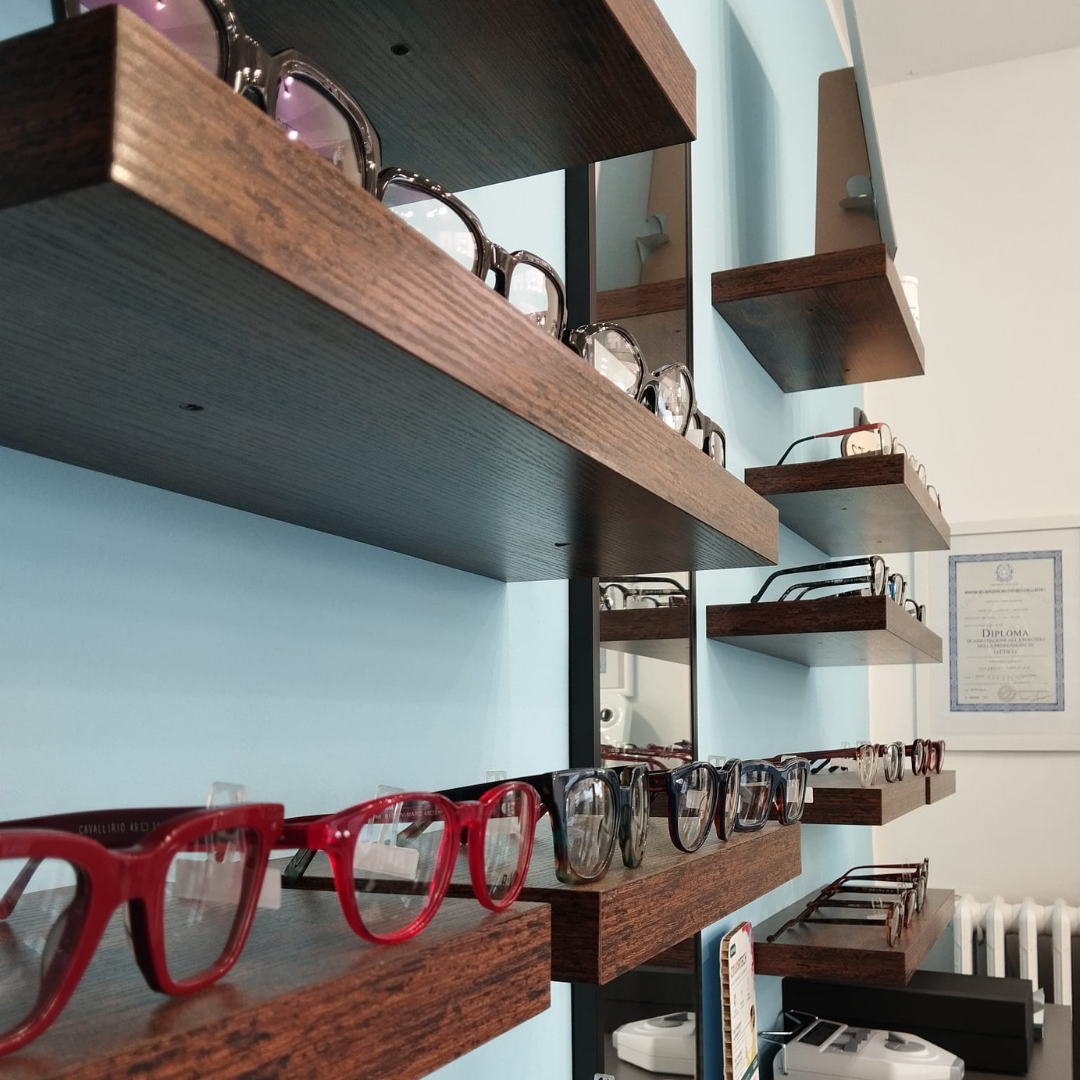 Occhiali da vista esposti, disposti su una parete bianca. Sono visibili montature di vari stili e colori.