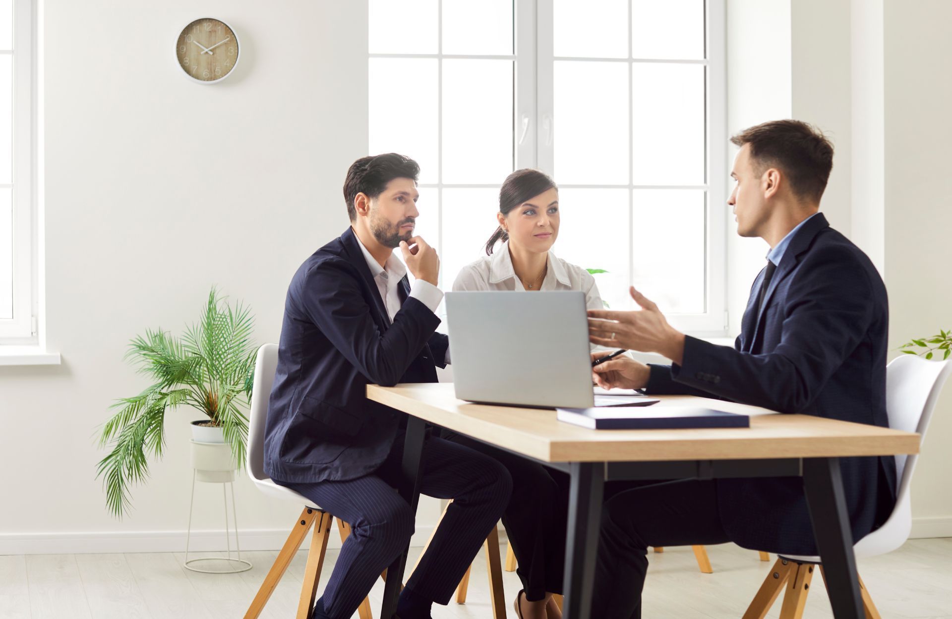 Three people in suits at a table: a man speaking, a man thinking, and a woman looking at a laptop in a bright office.