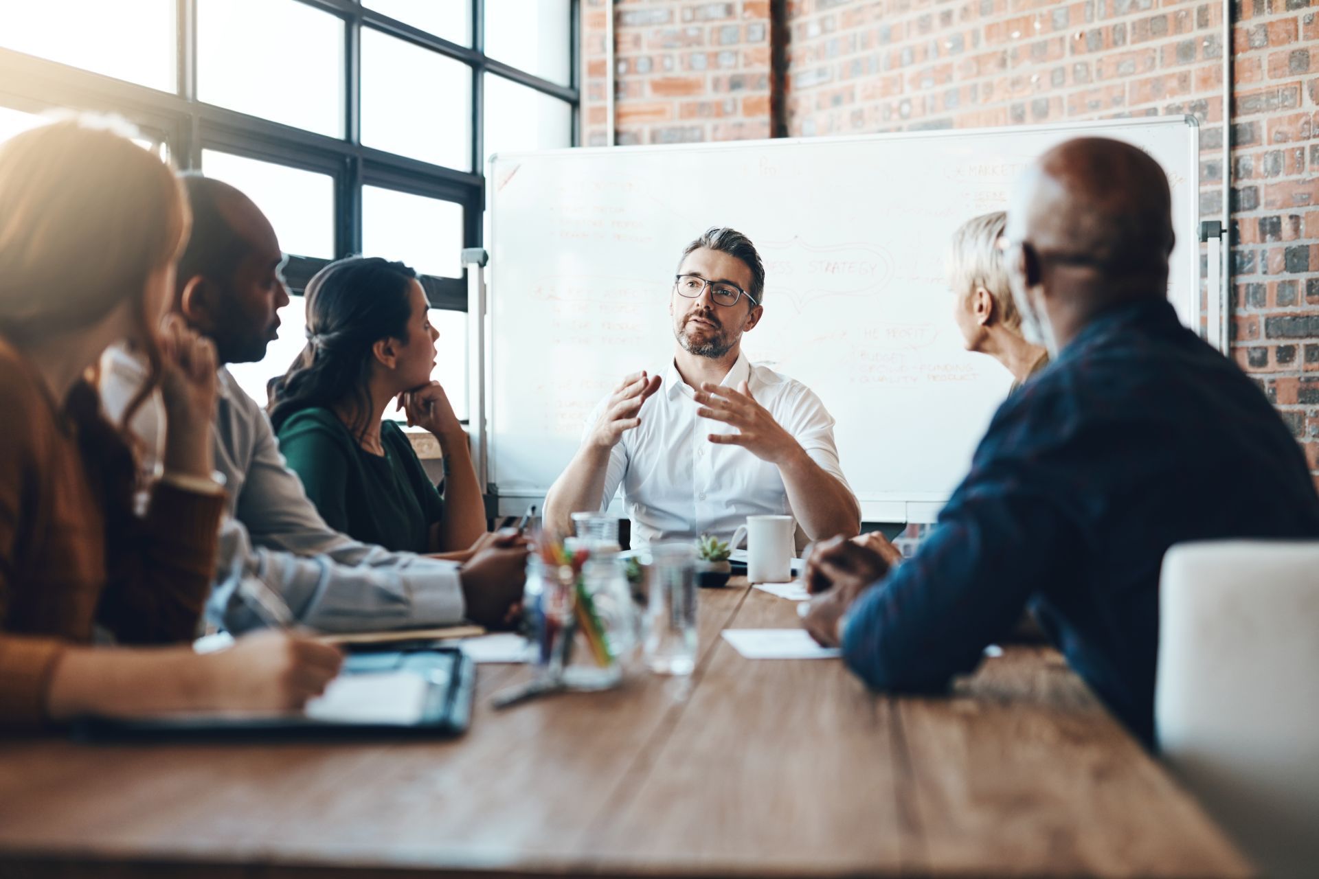 Business team in meeting, man gesturing by whiteboard, brick wall, wood table.