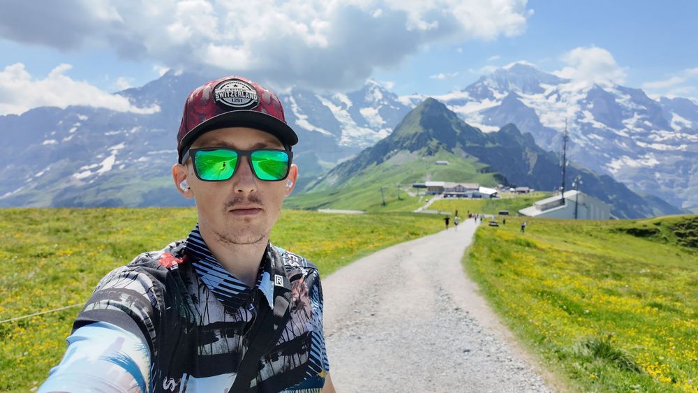 Selfie of a man in sunglasses on a mountain path with snowy peaks and green hills behind him