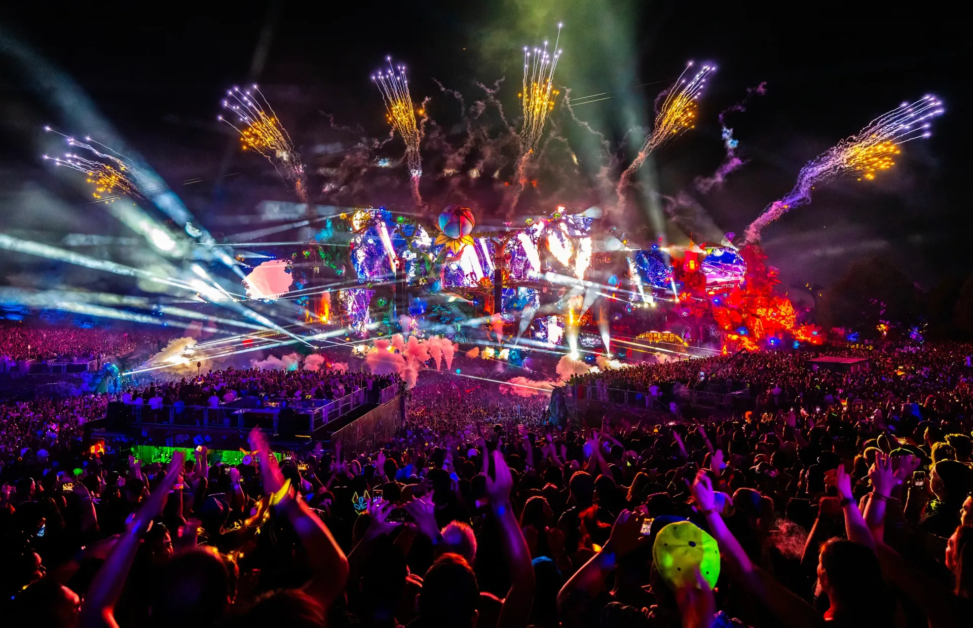 Crowd at a concert with colorful stage lights, fireworks, and raised hands at night