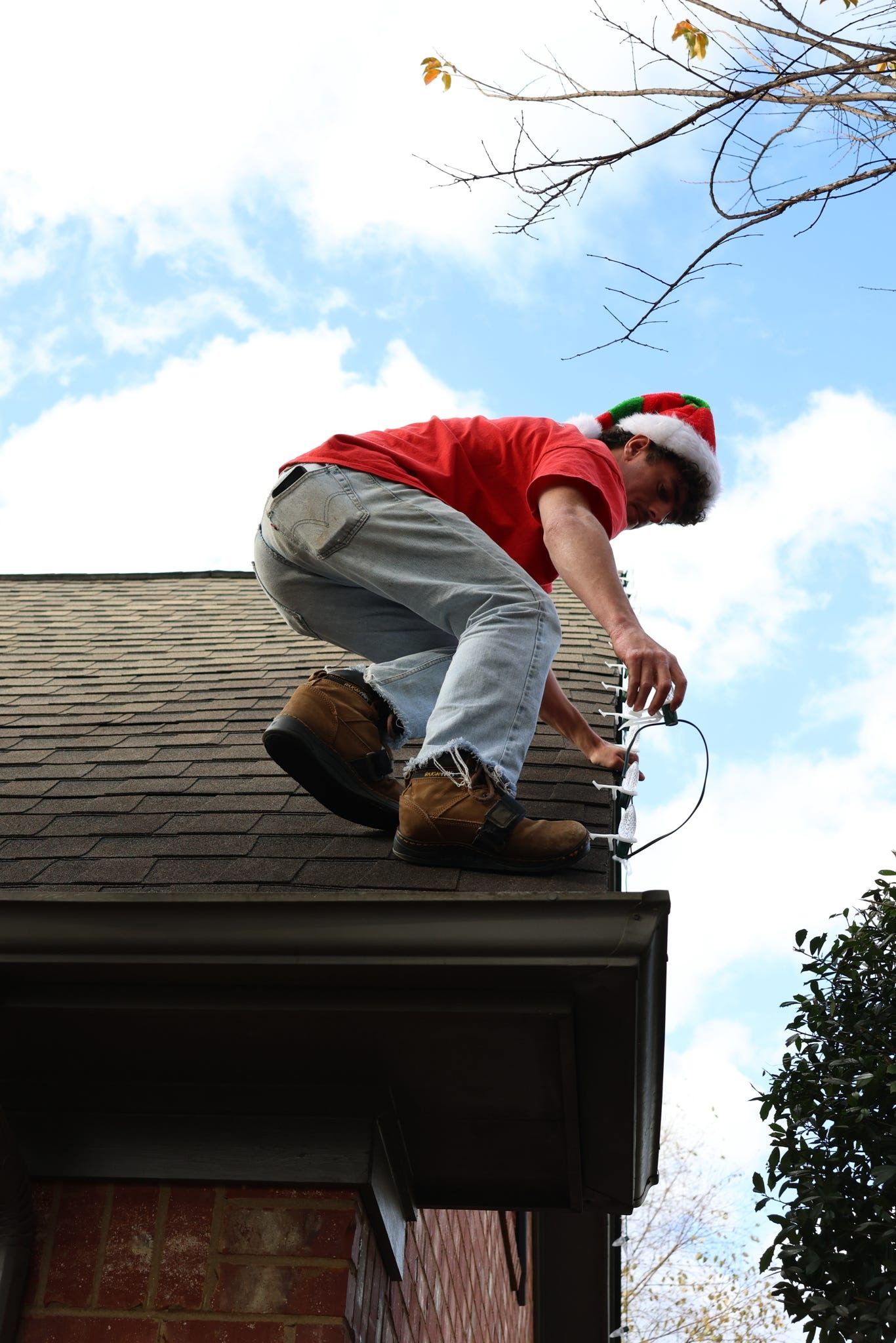 Man in Santa hat hangs Christmas lights from a roof gutter on a sunny day.