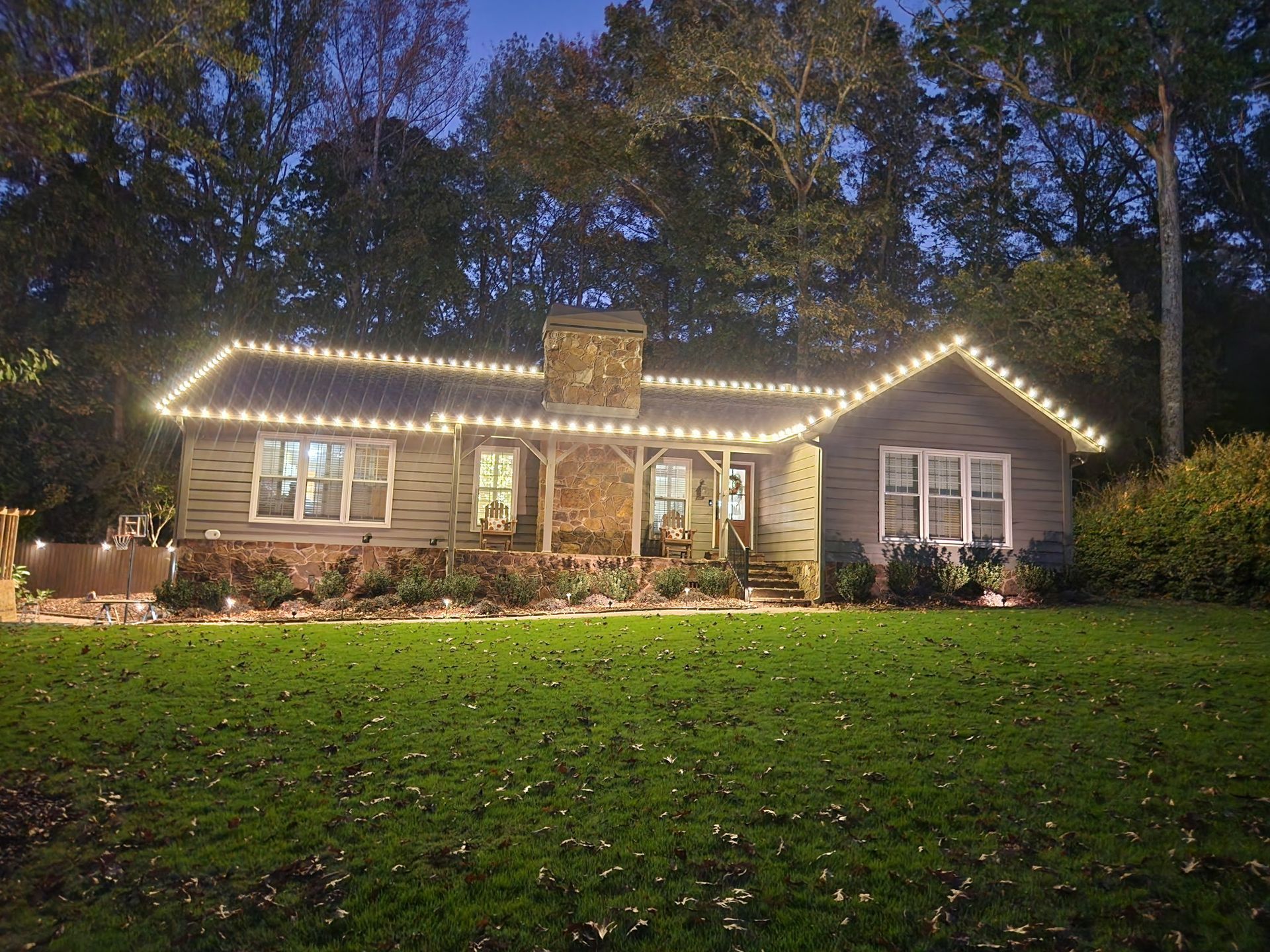 Cottage with Christmas lights on roof and porch, illuminated at dusk with green lawn.