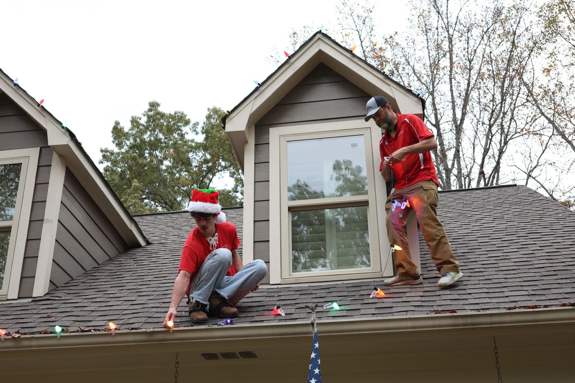 Two people on a roof hanging Christmas lights; one in a Santa hat.