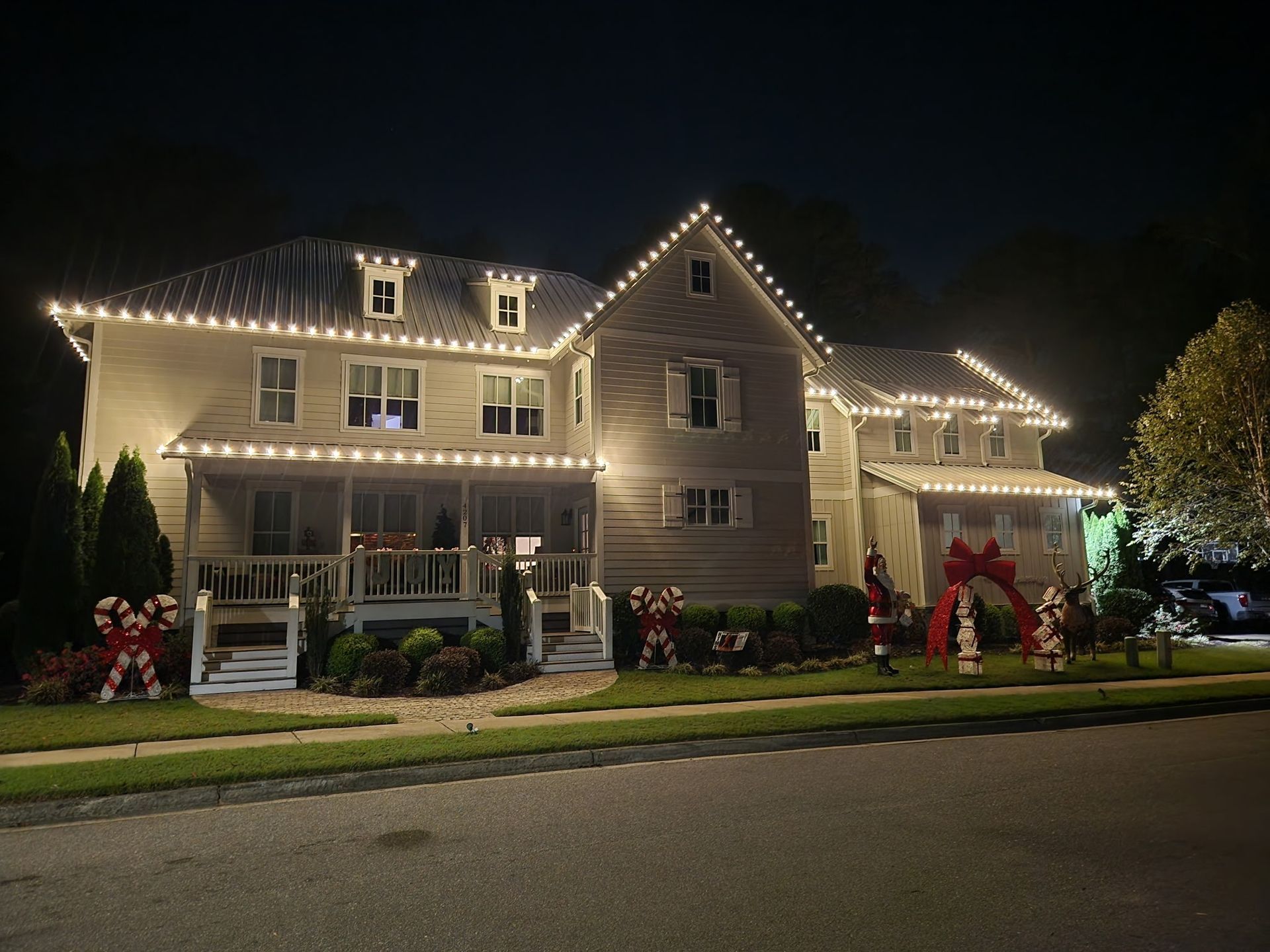 Large house decorated with white Christmas lights on roof and yard ornaments at night.