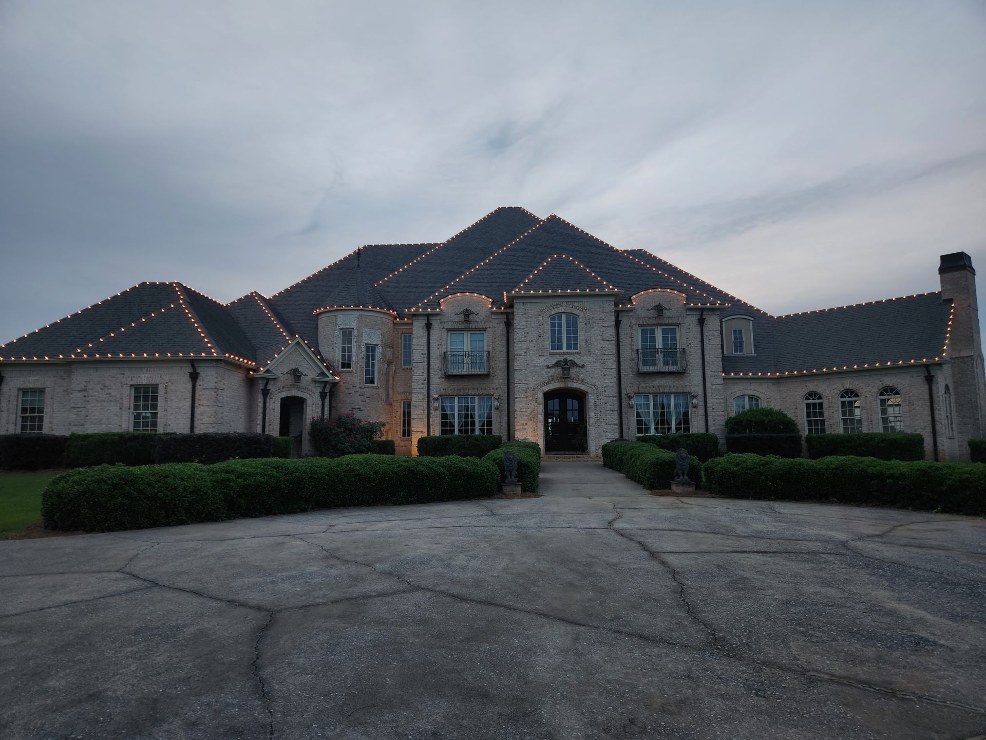 Large brick house with Christmas lights on the roof, under a cloudy sky.