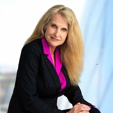 A woman in a black suit and pink shirt is sitting in front of a window.