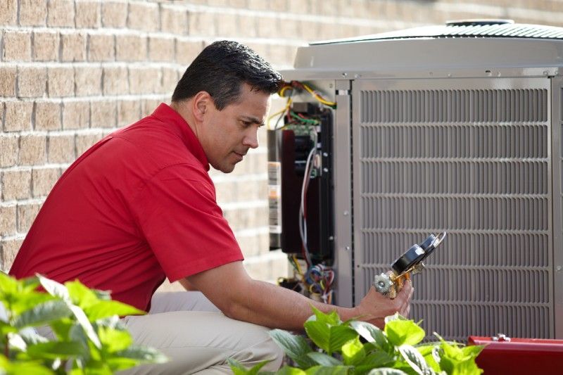 HVAC technician inspecting an air conditioning unit. Red shirt, brick wall, outdoor setting.