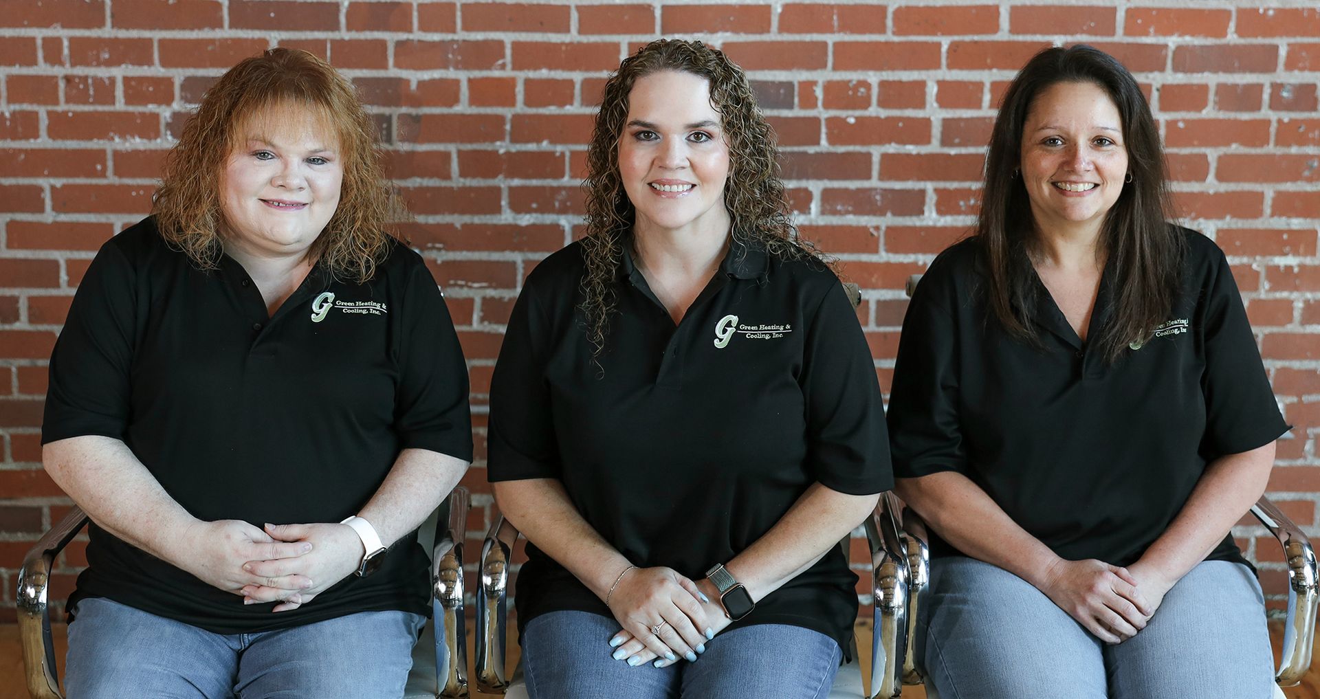 Three women in black shirts and jeans sitting in front of a brick wall, smiling.