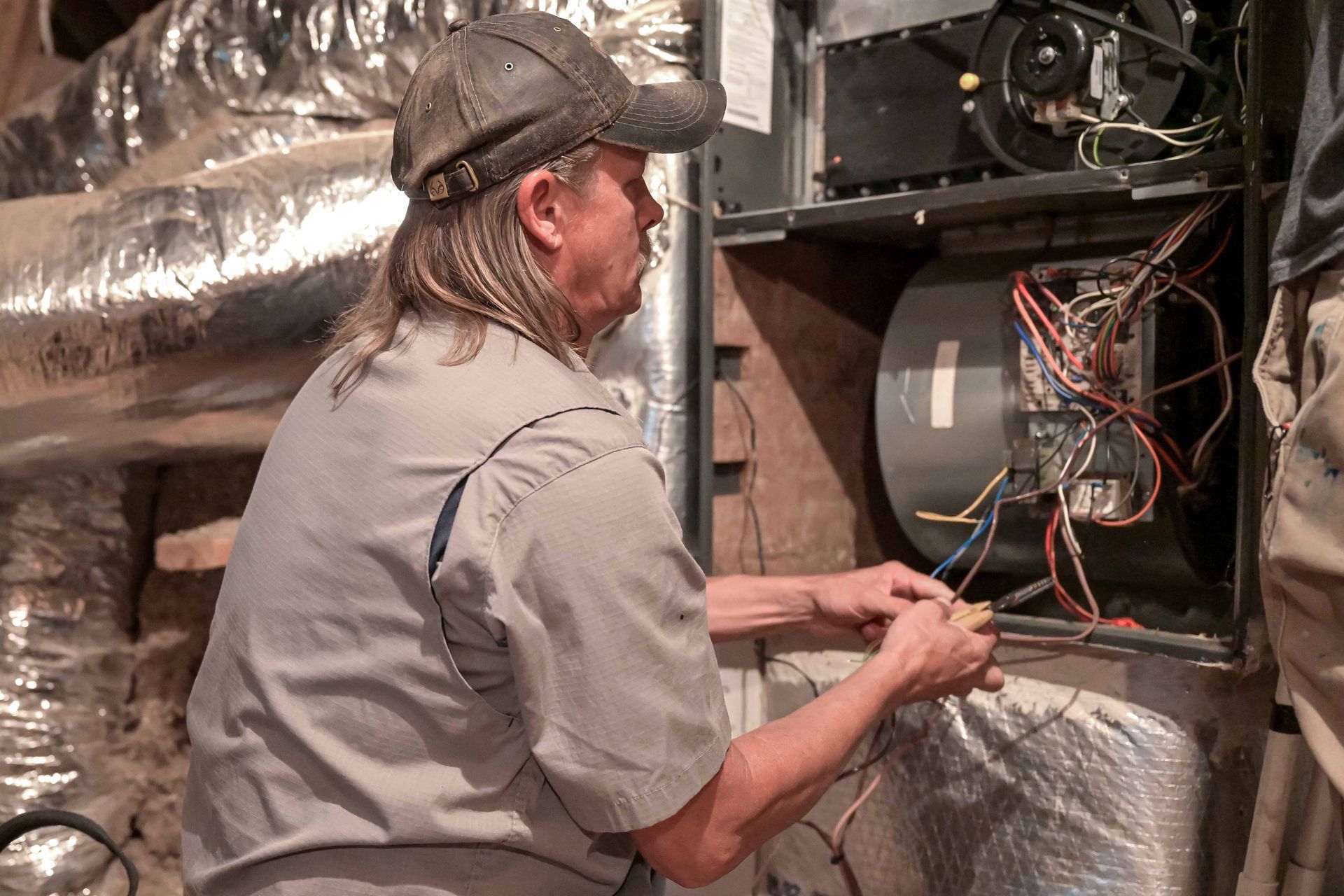 A technician working on electrical wiring inside an HVAC unit, wearing a cap.