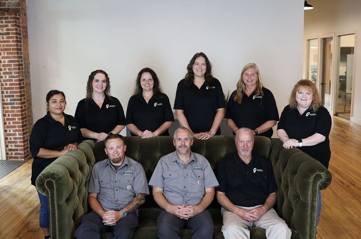 Group photo of men wearing gray work shirts, posing in front of a white wall, some seated on a green couch.