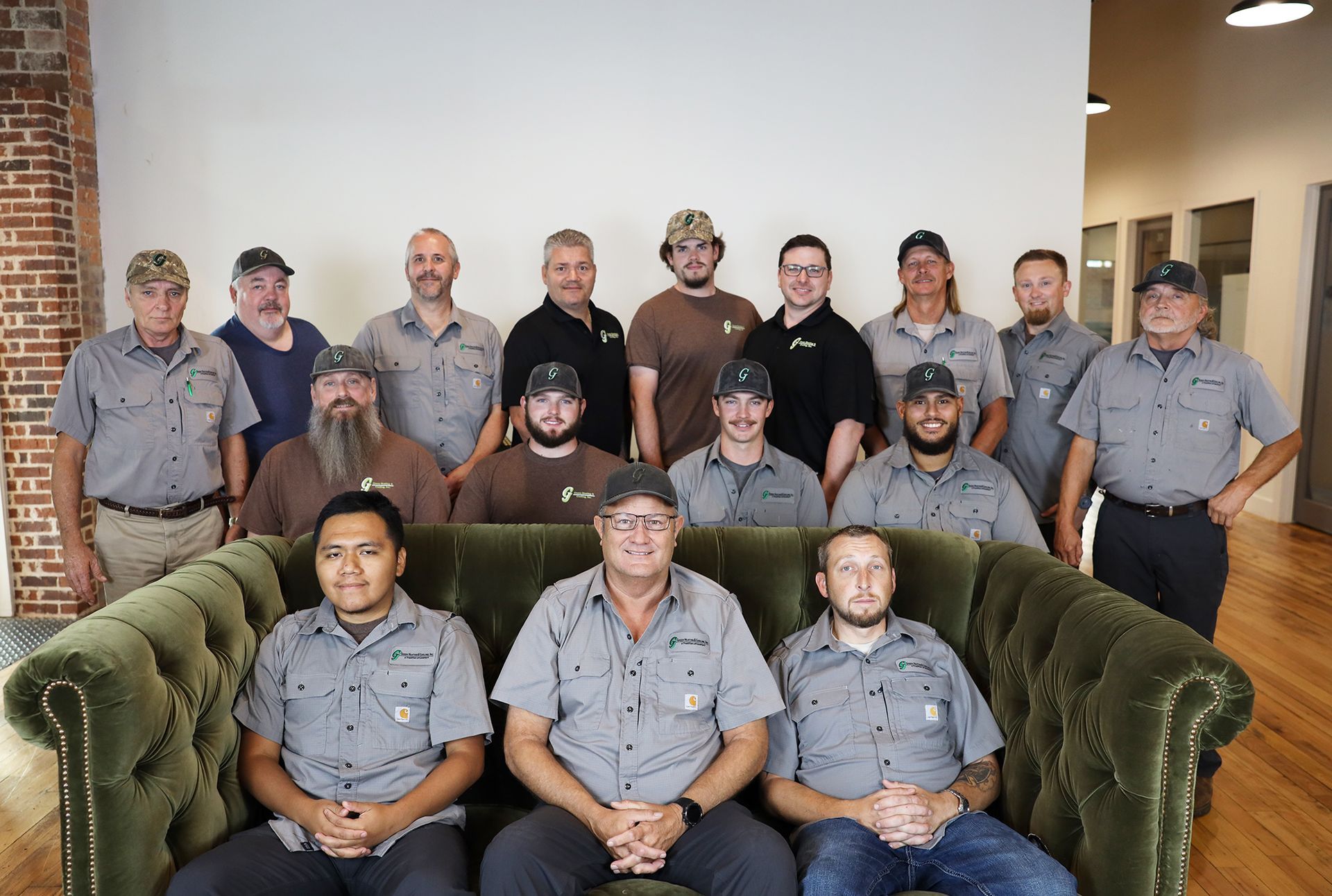 Group photo of men wearing gray work shirts, posing in front of a white wall, some seated on a green couch.