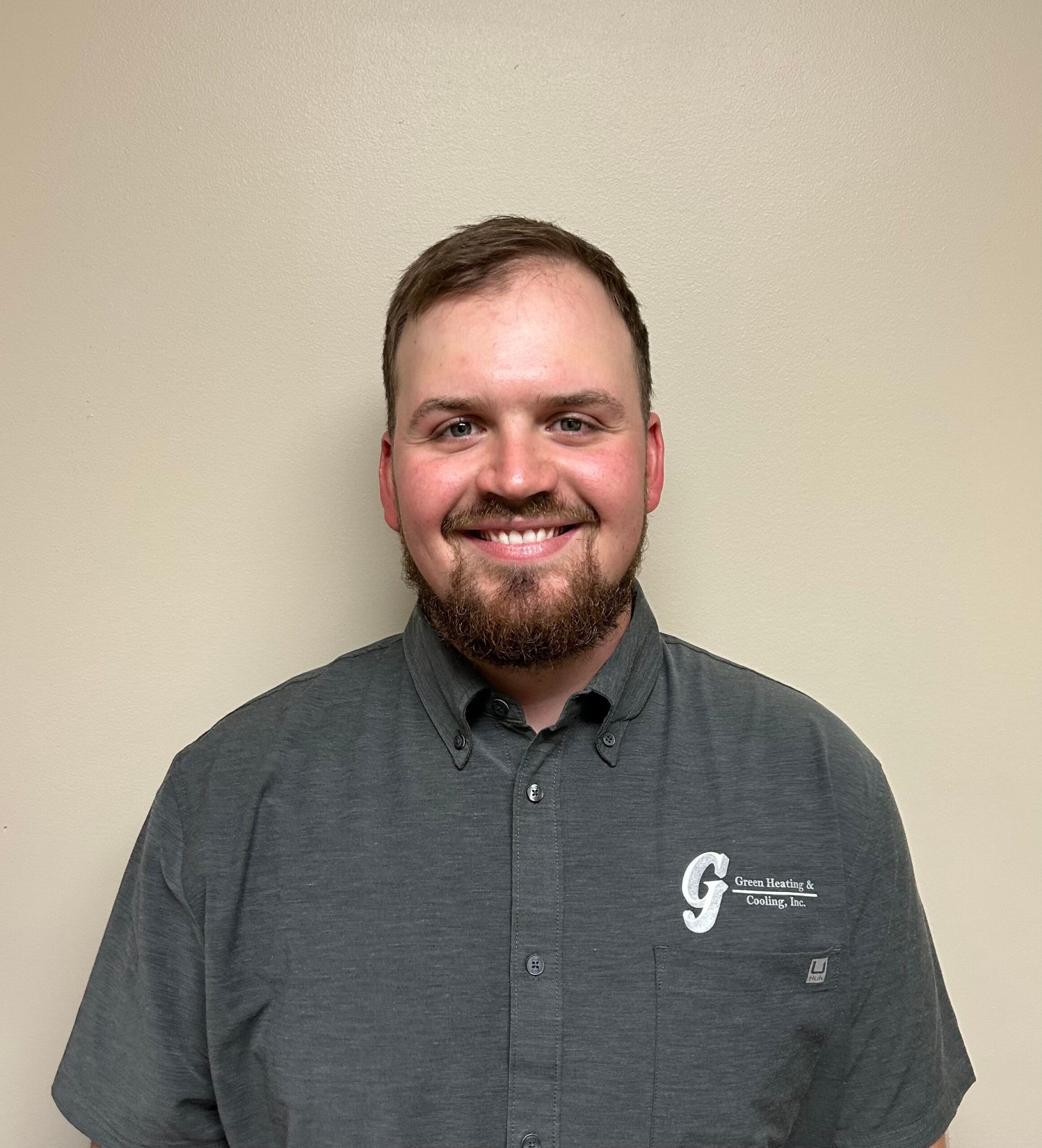 Man with a beard, wearing a gray shirt, smiling. The background is a plain, light wall.