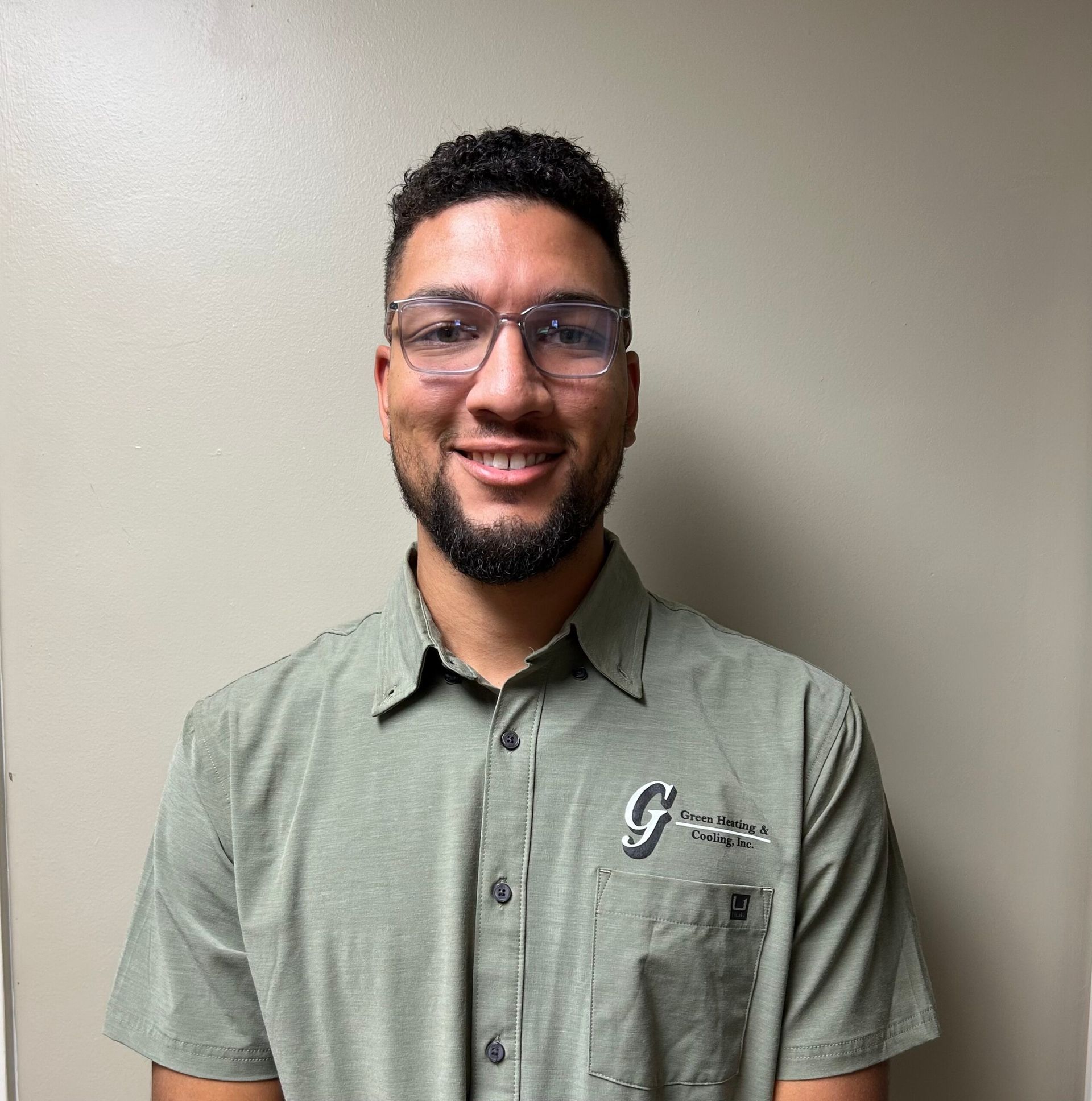 Person wearing glasses and a green collared shirt smiles at the camera. Neutral background.