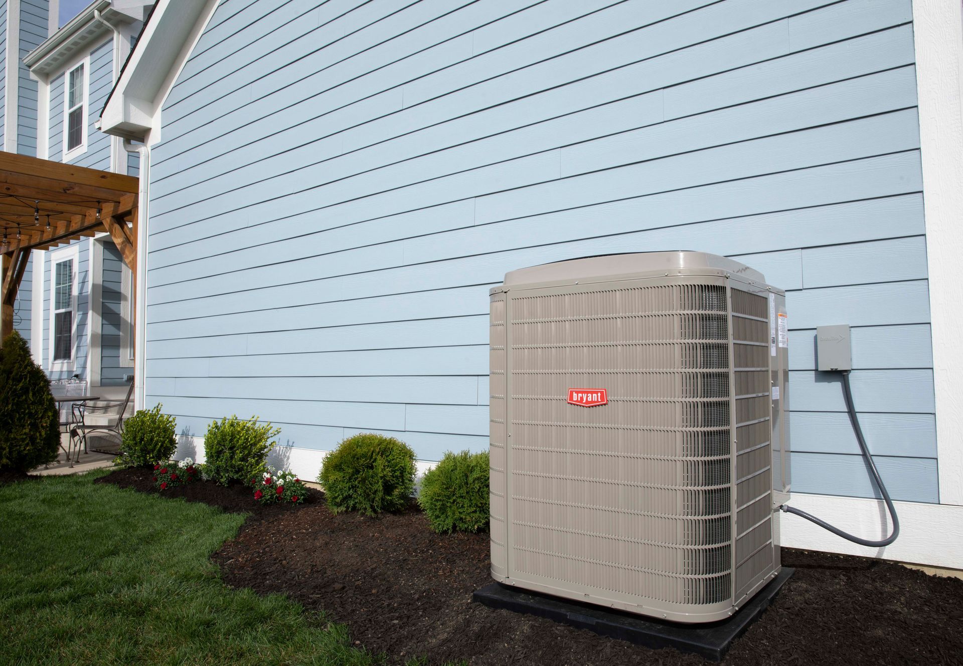 Air conditioner unit next to a light blue house on a green lawn with shrubs.
