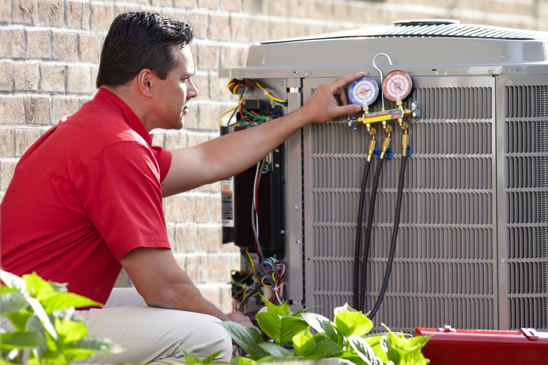 HVAC technician in red shirt, kneeling, attaching gauges to an outdoor air conditioning unit.