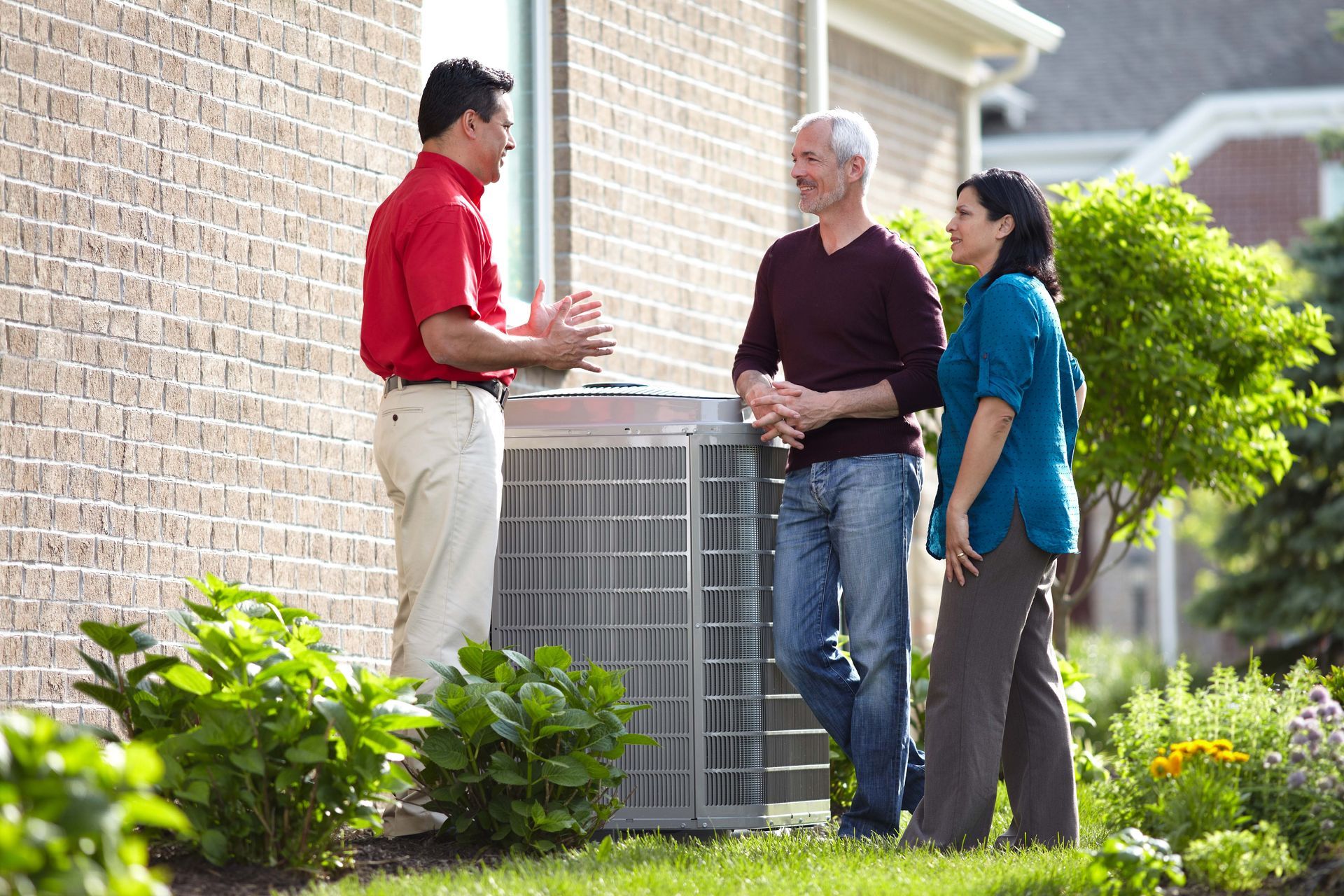 Man in red shirt discusses HVAC unit with a couple in a yard.
