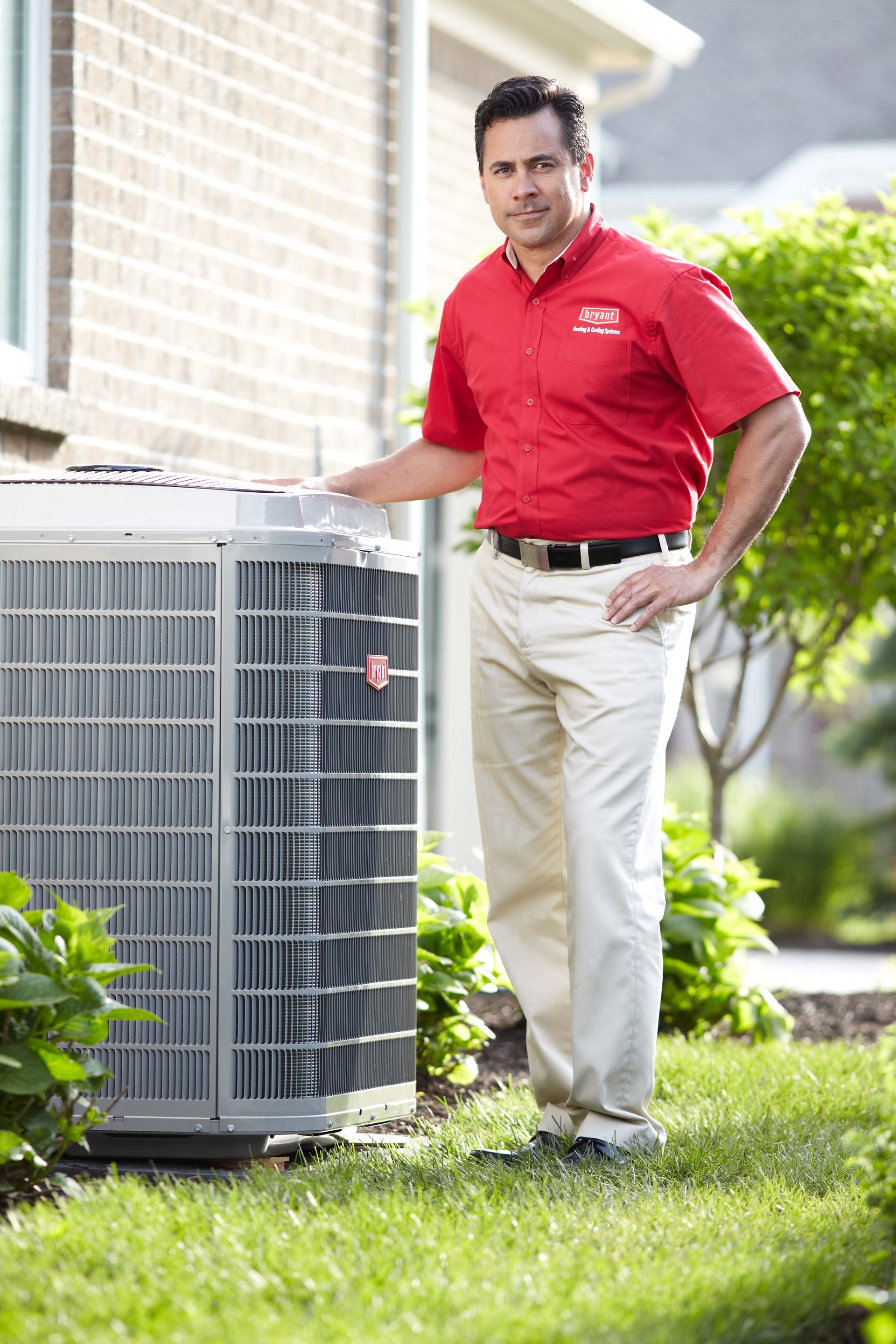 HVAC technician in red shirt and khakis beside an air conditioning unit outside a brick building.