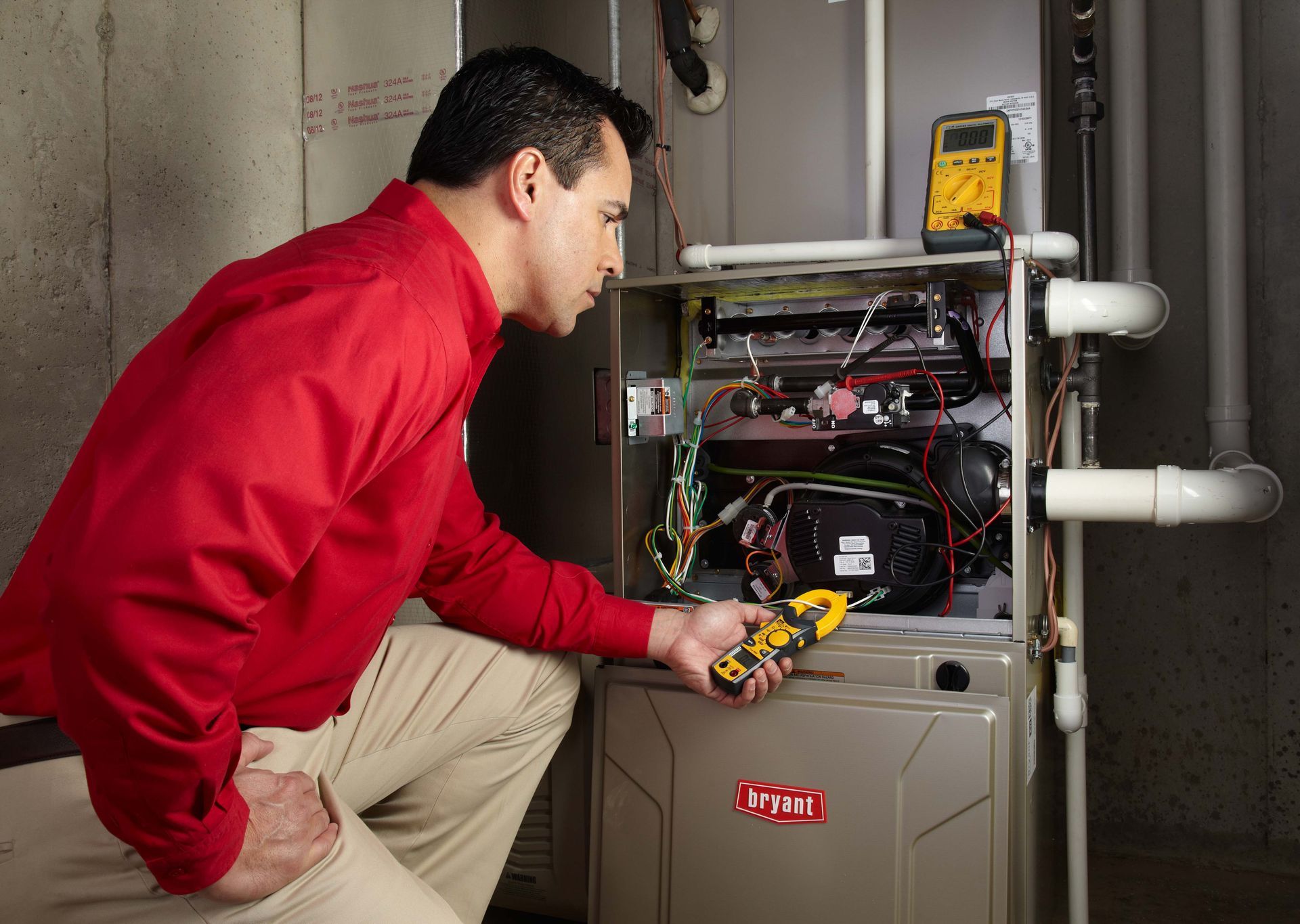 Man in red shirt kneels, checking furnace with a multimeter; gray furnace, white pipes, in a utility room.