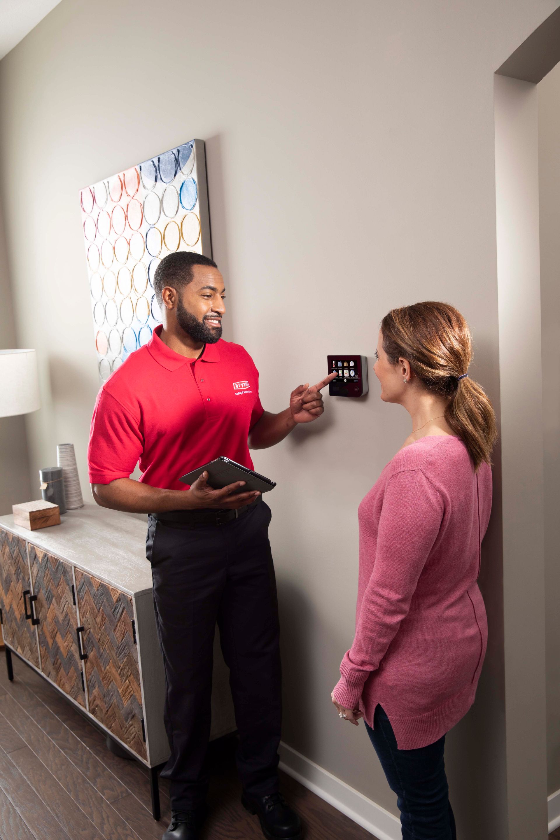 Man in red shirt points to a thermostat, talking to a woman. They stand near a console table with art.
