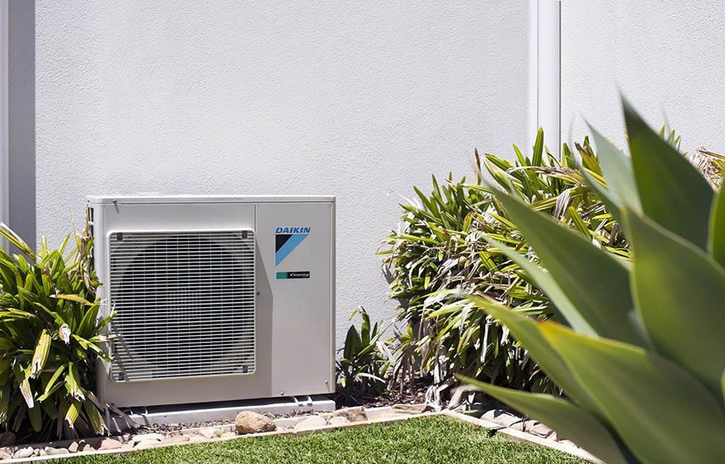 Air conditioner unit mounted outside, against a stucco wall, surrounded by greenery.