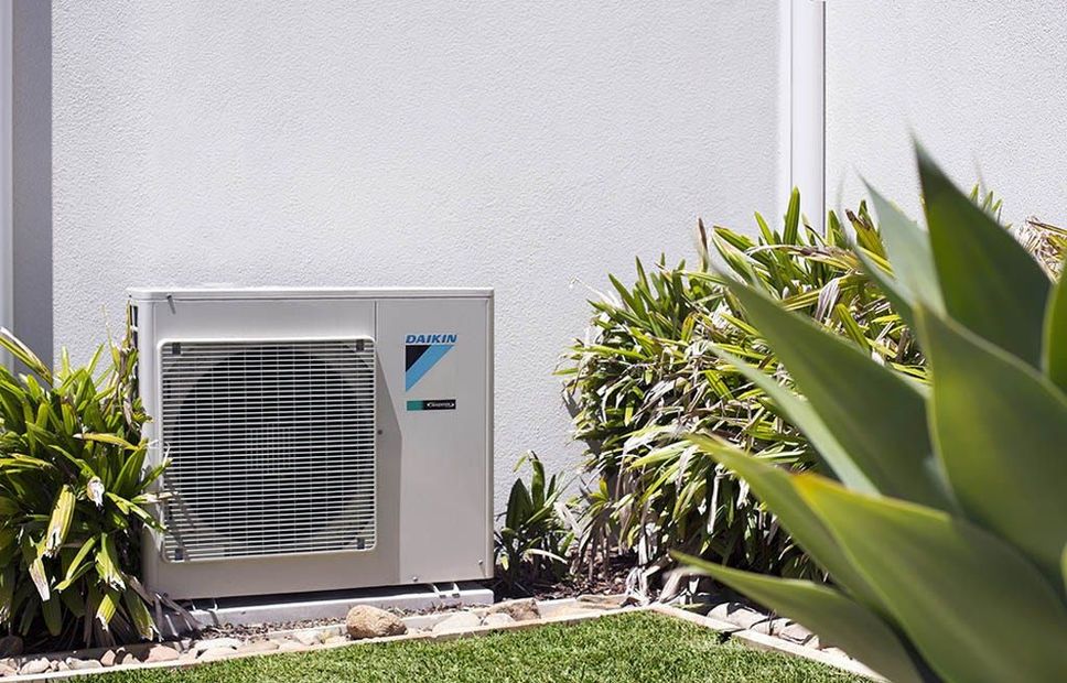 Air conditioner unit against a textured wall, surrounded by green plants and grass.