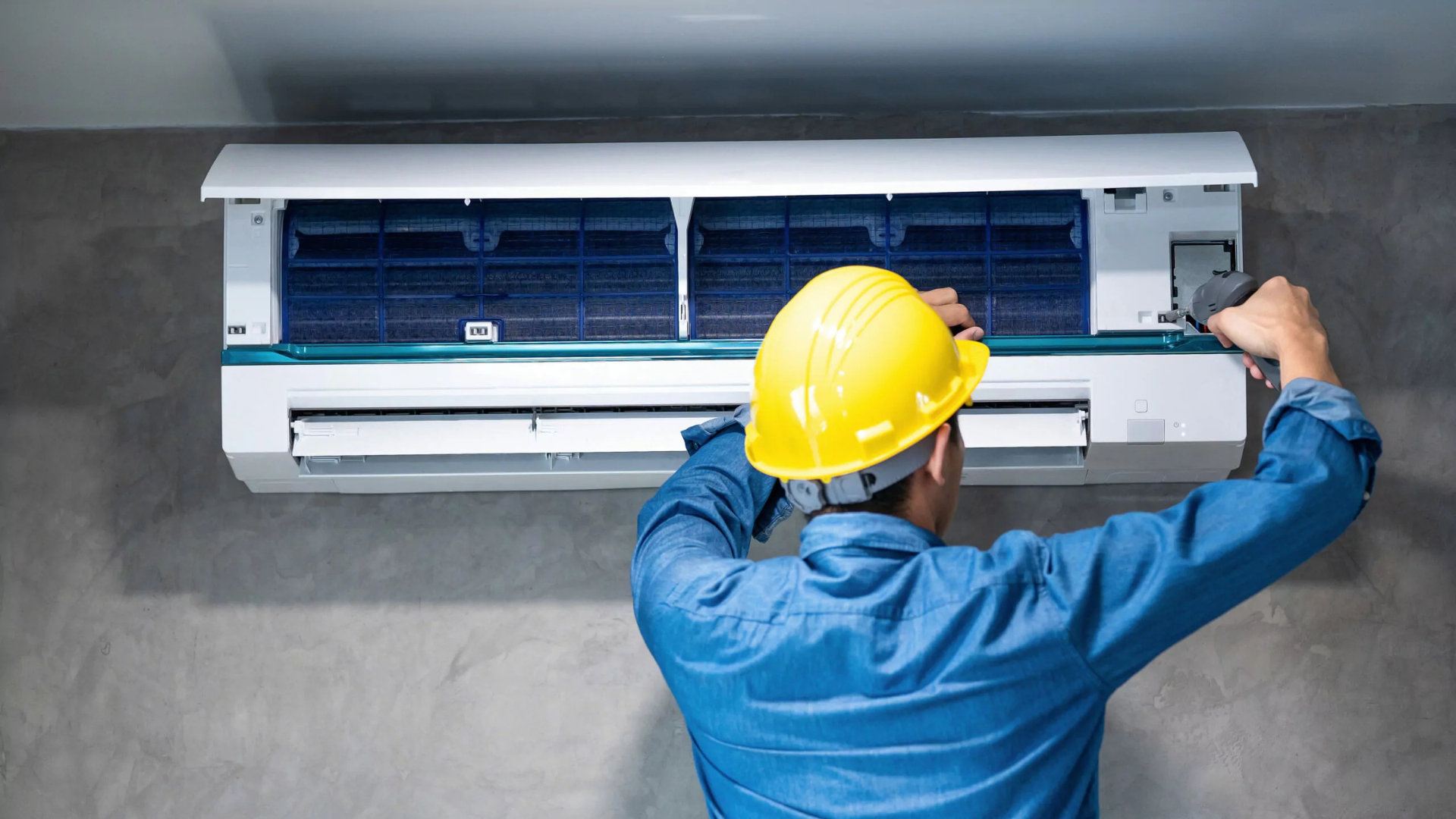 Technician in blue overalls and yellow hard hat servicing an air conditioning unit mounted on a gray wall.